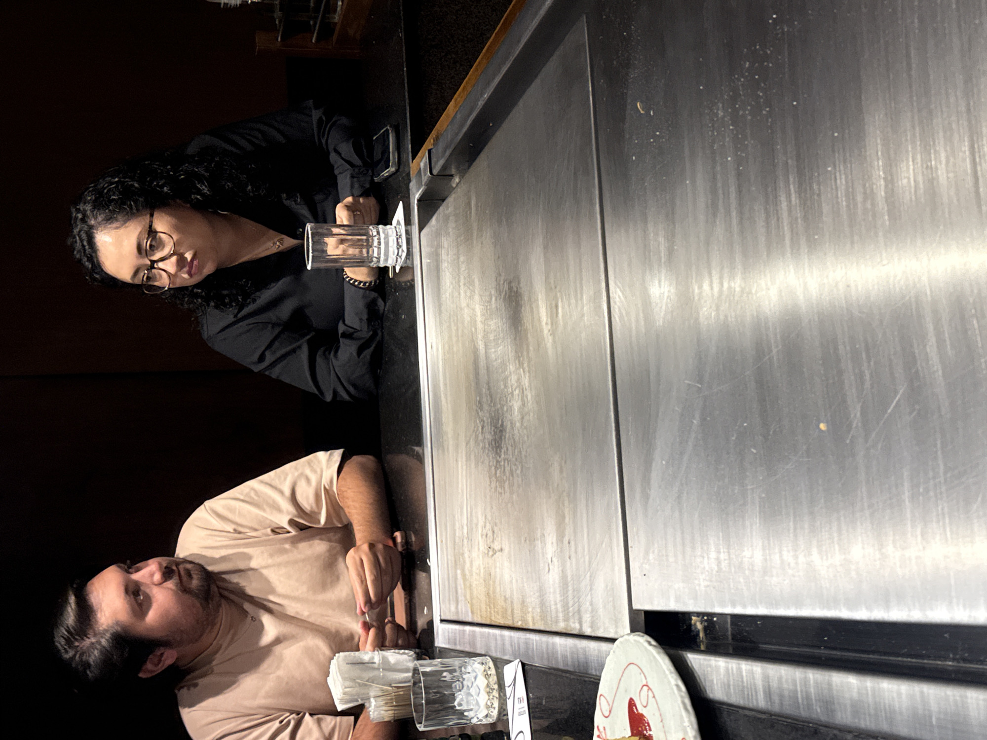 The image, rotated 90 degrees counter-clockwise, depicts two individuals seated at a teppanyaki-style restaurant, characterized by a large, reflective stainless steel cooking griddle that dominates the right side of the frame. The overall lighting is dim, suggesting an evening setting.
On the left, a woman with dark, curly hair and glasses, wearing a black long-sleeved top, is seated. She looks towards the viewer with an observant expression, holding a tall, clear glass. Her phone is visible on the dark countertop next to her.
Below her, a man with a beard and dark hair, dressed in a light tan t-shirt, is also seated. He looks slightly upwards and to his right, appearing relaxed. His hands are resting near a stack of white napkins and another clear glass on the counter. A portion of a white plate with red patterns is visible in the bottom right corner.
The extensive metal griddle shows signs of wear, including scratches, minor residue, and some discolored areas, indicating past use for cooking. The individuals appear to be diners, likely observing or waiting for food to be prepared on the griddle. A small, white card near the man's glass has partially visible text, including the word "THE". The scene is an indoor restaurant setting, consistent with a dining experience in Puebla, Mexico.