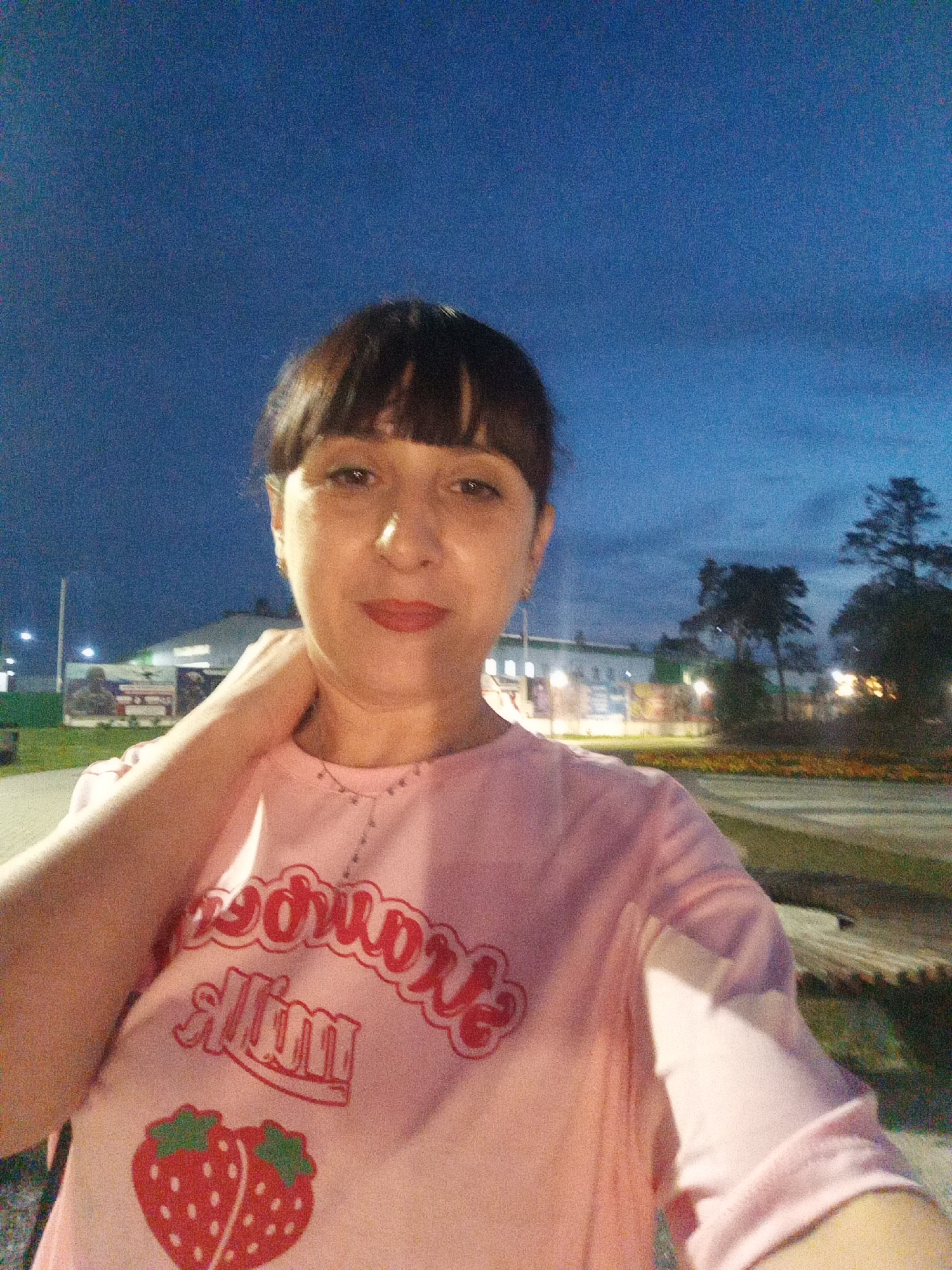 A woman takes a selfie outdoors at dusk in Belogorsk, Russia. She is wearing a pink t-shirt with a strawberry graphic and text that reads "Strawberry Milk." The background shows a park with trees and buildings illuminated by artificial lights. The sky is a deep blue with visible clouds.