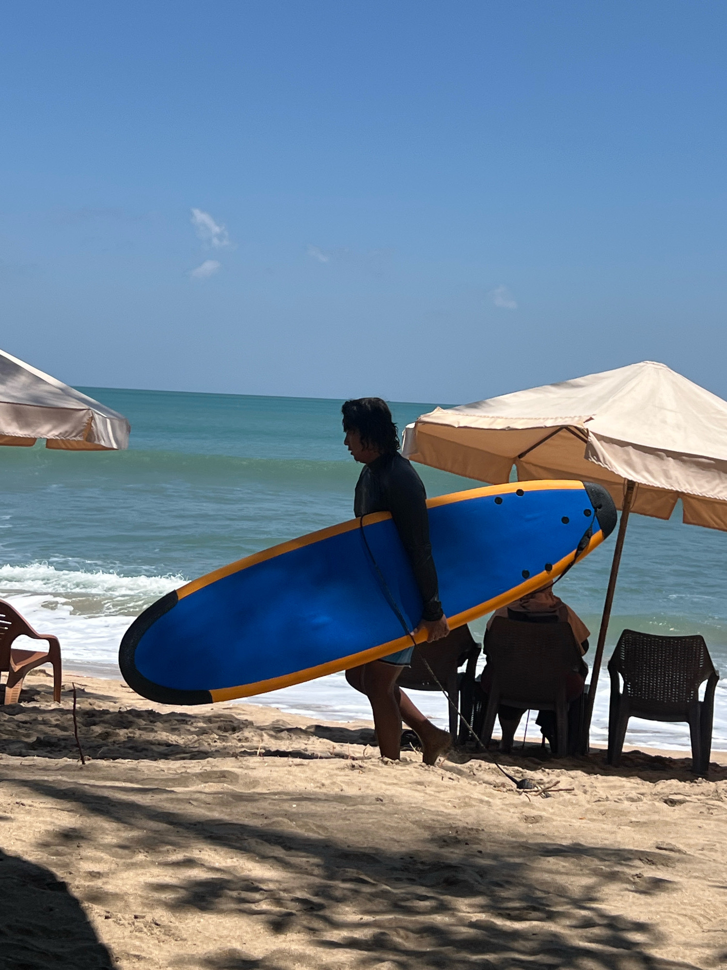 A person is walking on a sandy beach in Kuta, Indonesia, carrying a blue and orange surfboard. The person is wearing a dark long-sleeved shirt and blue shorts. The ocean is to the left, with waves breaking on the shore. Two beach umbrellas provide shade for two people sitting in chairs. The sky is clear and blue with a few wispy clouds. The sand is light brown and marked with shadows from the umbrellas and trees.