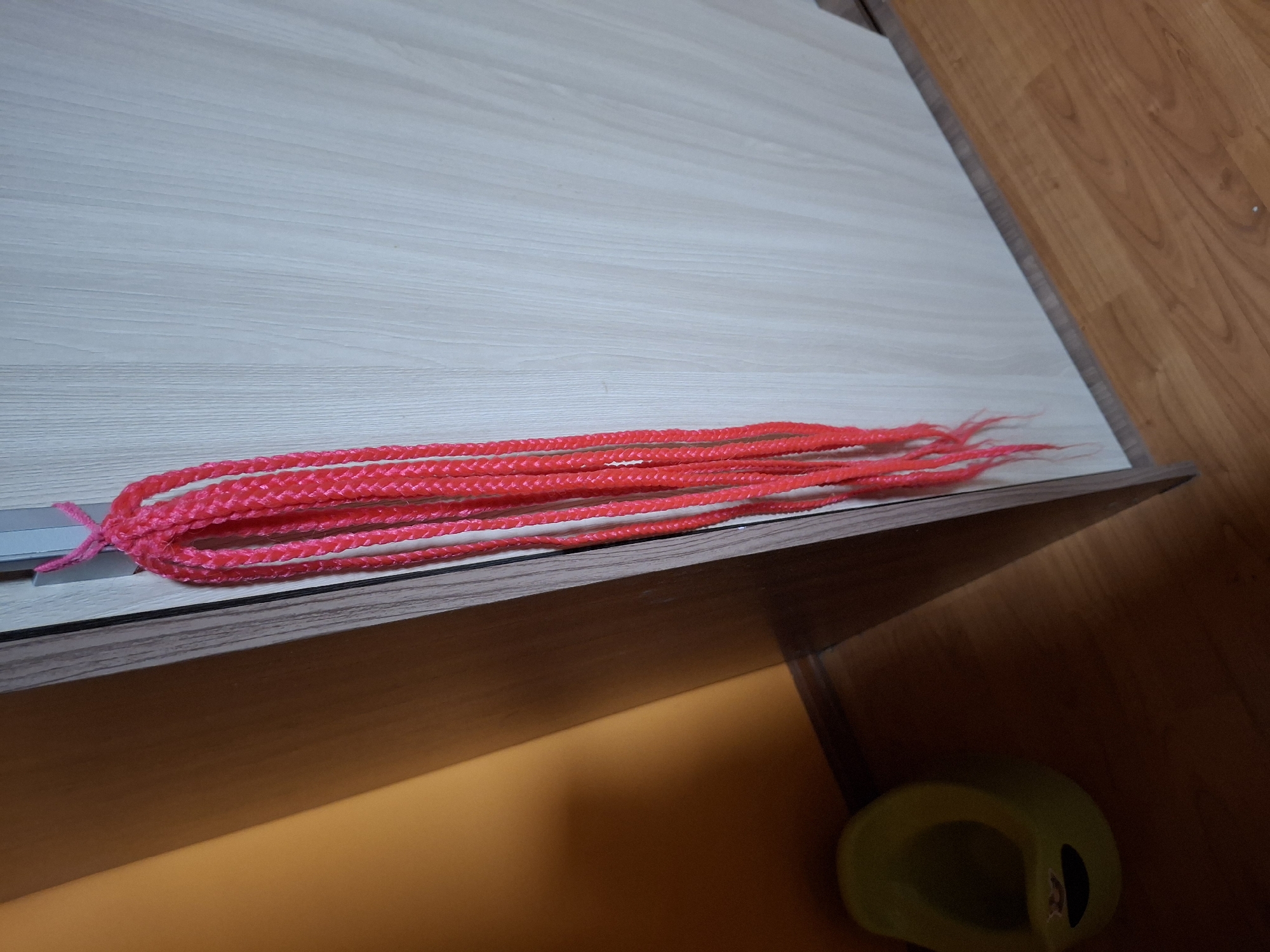 A close-up shot displays several bright pink braided strands of what appear to be synthetic hair or yarn, laid out on a light-colored, wood-grained surface. The braids are neatly done and run horizontally across the frame, with a pink tie holding a section of them together on the left side. The surface they rest on has a subtle texture, hinting at a laminate or veneer. To the right of this surface, a darker, polished wood floor is visible, with a distinct grain pattern. The scene is illuminated by warm light, casting a soft shadow below the surface where the braids are placed, suggesting the light source is from above and slightly to the left. The overall ambiance is neutral and simple, focusing on the texture and color of the braided material against the backdrop of the surfaces. There is a yellow object, possibly a plastic holder or cap, visible in the lower right corner of the frame, partially in shadow. No people are visible in the image. The setting appears to be indoors, likely a room with furniture and flooring.