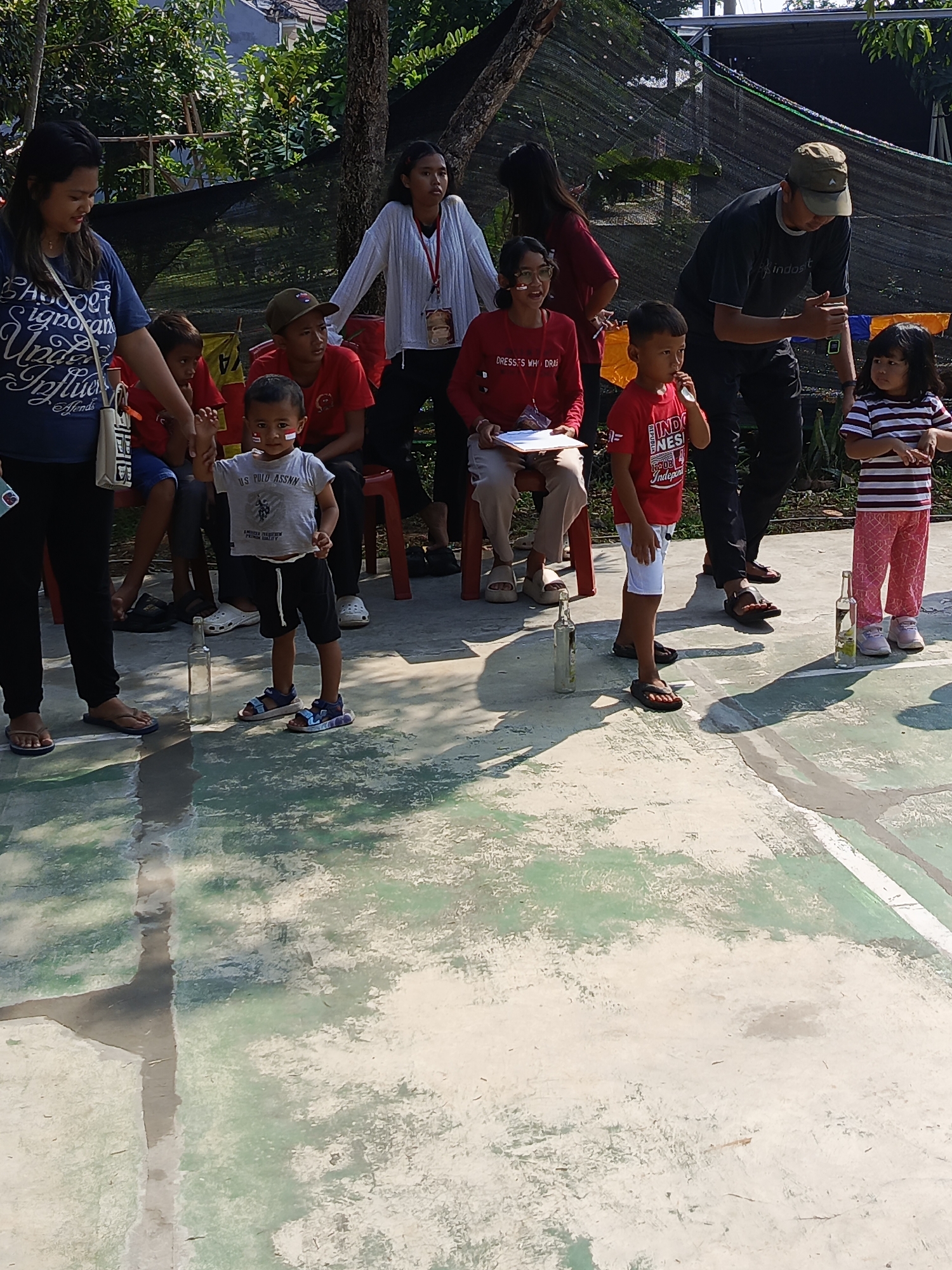 In the city of Ciseeng, Indonesia, on a sunlit day, a group of people, primarily children and adults, are participating in an outdoor event. The scene unfolds on a weathered concrete surface, possibly a sports court, marked with faded green paint and white lines.
A young boy, standing near the center of the frame, is the focal point. He is wearing a gray t-shirt with the text "US POLO ASSN." and black shorts, accompanied by blue and black sandals. His right hand is raised, seemingly engaged in a game. Notably, a small Indonesian flag is painted on his left cheek.
To his left, a woman in a blue t-shirt with decorative lettering and black pants stands, looking down at the boy. Her left hand is extended towards him, possibly guiding or encouraging him. Beside her, a younger boy in a red t-shirt is partially visible.
To the boy's right, another child, a boy in a red t-shirt with white writing that includes "INDONESIA" and "INDEPENDENCE", stands with a white object in his mouth. He is wearing white shorts and dark flip-flops.
Further to the right, a young girl with long dark hair, dressed in a red and white striped t-shirt and patterned pink pants, stands with her hands by her side, observing the activity. She is wearing white sneakers.
An adult male, wearing a dark t-shirt, black pants, and a green cap, is also present, positioned to the right of the children and appearing to be engaged in the game, possibly preparing to place or retrieve an object.
Several glass bottles are strategically placed on the ground, suggesting a game involving targets or challenges. In the background, lush green trees and foliage create a natural backdrop, with a black net structure visible above. The overall atmosphere appears to be a lively community event.