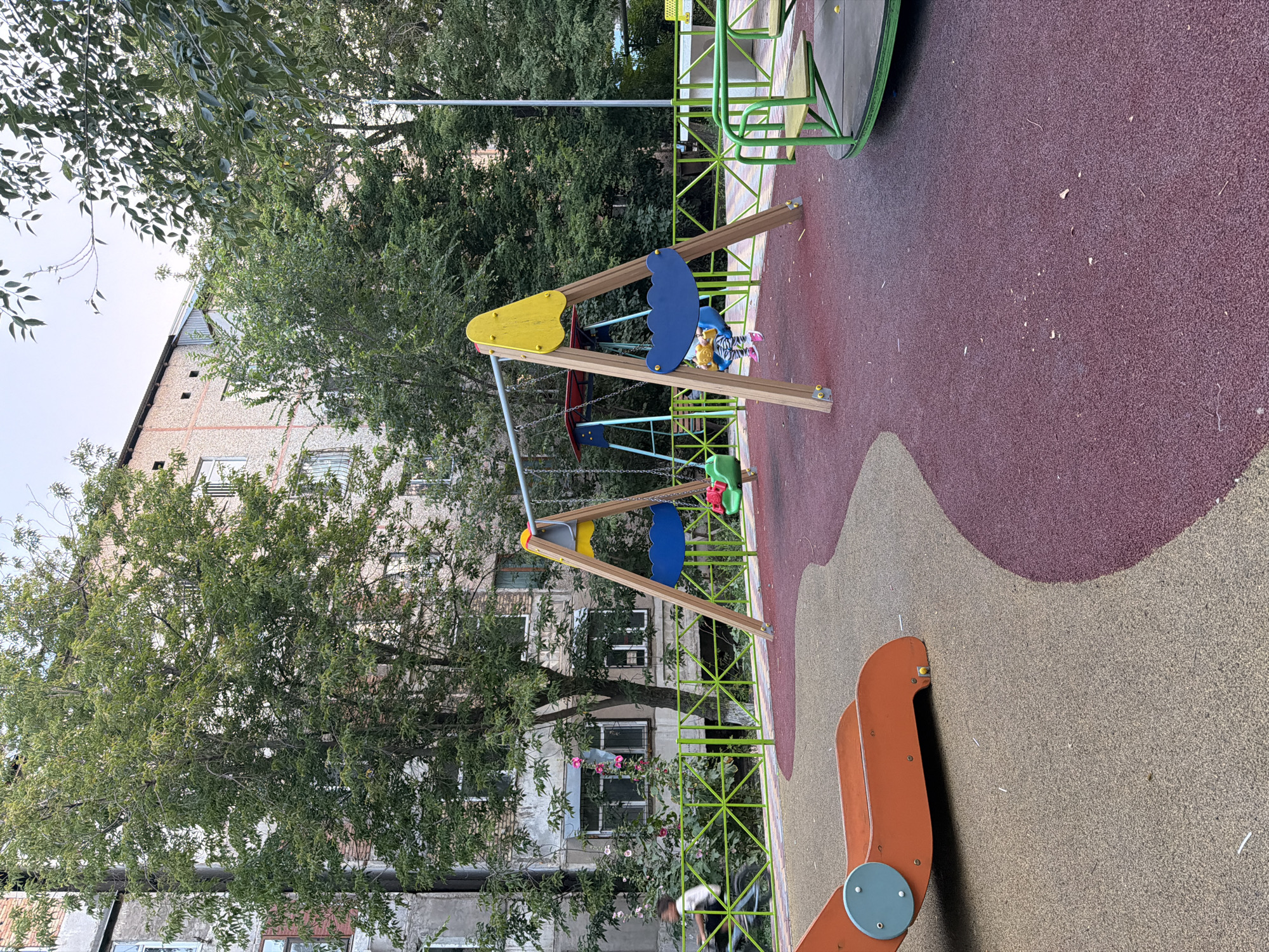 This is a high-angle, full shot of a playground with a building and trees in the background. The playground has a soft, rubberized surface in two colors: a reddish-purple and a light tan. In the foreground, part of an orange seesaw-like structure is visible, with a light blue circular cap on one of its supports.
Further into the playground, a swing set with a wooden A-frame structure is prominent. One of the swings has a yellow seat and a blue curved back, and it appears to be occupied by a child wearing striped clothing and pink shoes. The child is leaning forward, suggesting they are actively swinging. Another swing, partially obscured, is also visible. A green metal fence encloses part of the playground.
Behind the playground, a multi-story building with a textured facade and numerous windows can be seen. Large, leafy trees with dense foliage partially obscure the building and extend across the upper portion of the frame. Some small pink flowers are visible in the vegetation near the building.
The lighting suggests it is daytime, possibly late afternoon, given the angle of the light and the overall brightness. The sky is visible in patches through the trees and appears overcast or hazy. There is no visible text in the image. The scene conveys a sense of everyday activity at a local playground in Shymkent, Kazakhstan.