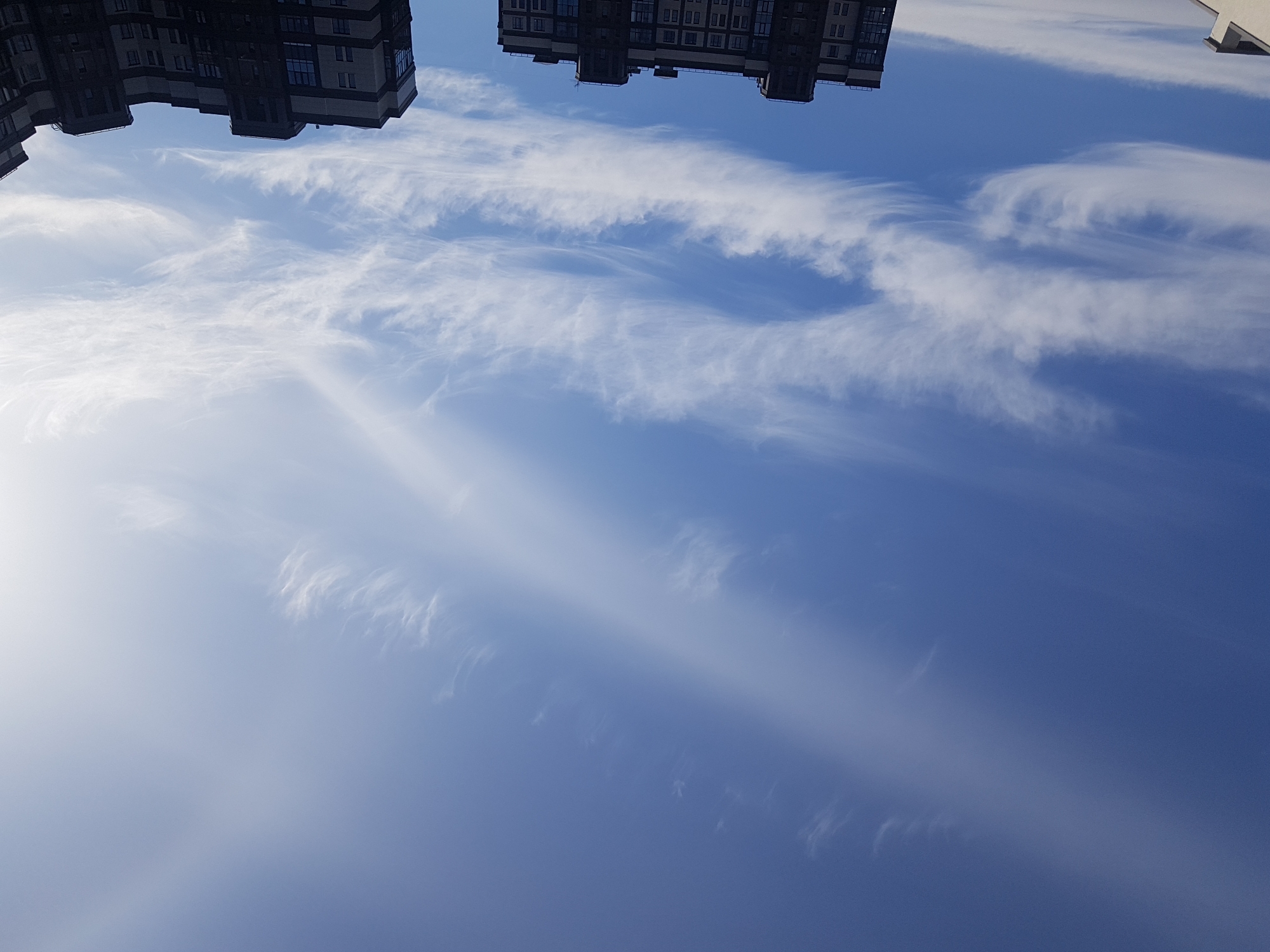 The image shows a view of the sky above Saint Petersburg, Russia. Wispy cirrus clouds are spread across a blue sky. Two buildings are visible at the top of the frame, upside down. The buildings are residential and appear modern. The sun is visible on the left side of the frame. The lighting suggests it is daytime.