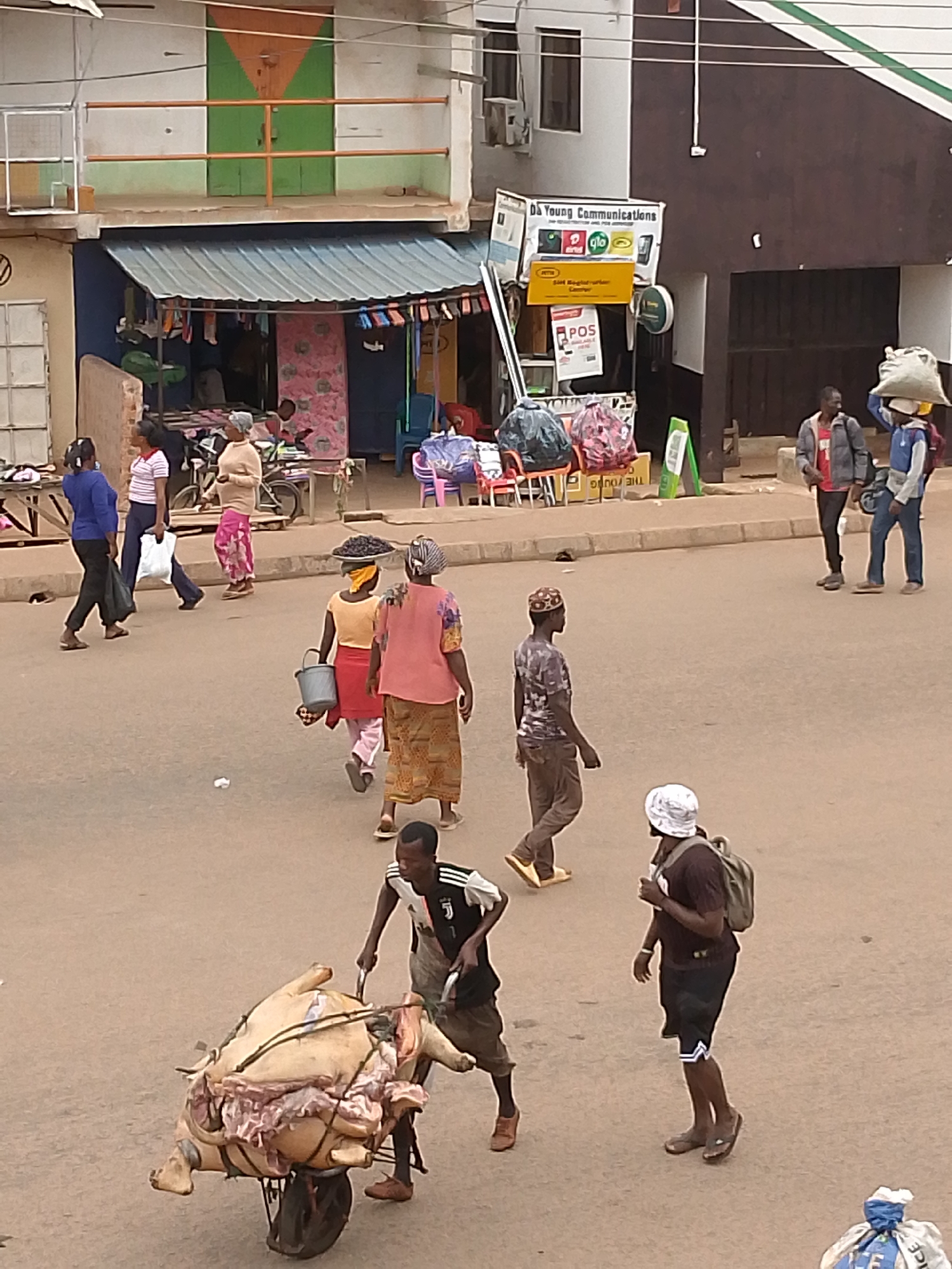 The image depicts a street scene in Bukuru, Nigeria. The road is unpaved and crowded with pedestrians. A man is pushing a wheelbarrow loaded with a large portion of raw meat, possibly pork. Other people are walking by, some carrying items such as buckets or bags on their heads. In the background are buildings with various businesses, including one with a sign that says "Da Young Communications." There are also stalls or shops selling goods under awnings. The overall scene shows a busy and typical day in an urban area.