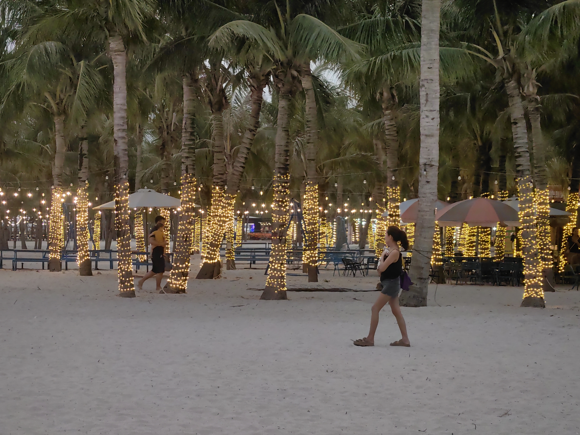 The image shows a beach scene at dusk in Bai Chay Ward, Vietnam. Tall, slender palm trees line the beach, their trunks wrapped with strings of yellow fairy lights. The lights illuminate the sandy ground, creating a warm glow. People are walking on the beach. A woman wearing a dark top and shorts walks from left to right across the foreground. Another person in a yellow shirt and dark shorts walks in the opposite direction. In the background, benches and tables with umbrellas can be seen. The sky is overcast.