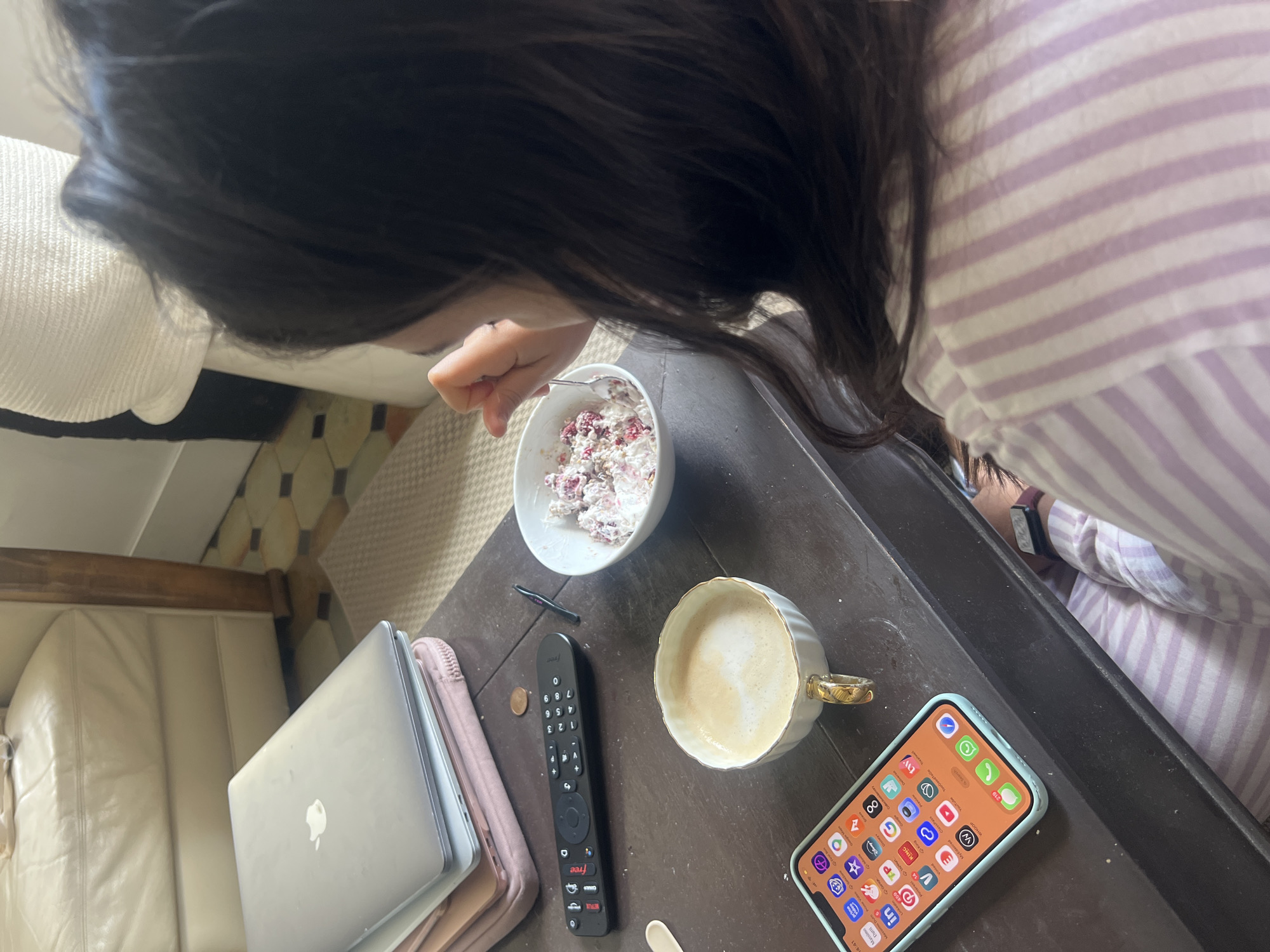 A woman is pictured from an overhead perspective, appearing to eat from a white bowl containing what looks like a yogurt or oatmeal-based mixture with red berries. The bowl is placed on a dark brown coffee table. To the left of the bowl, there is a silver laptop sitting atop what looks like a pink book or pad. Next to the laptop is a black remote control, along with a coin and a small item, possibly a pen or tweezers. A cup of coffee with milk is also on the table. An iPhone in a light blue case is placed near the cup. A portion of a light-colored couch is visible to the left, and striped clothing is seen on the person. The location cannot be determined from this image.