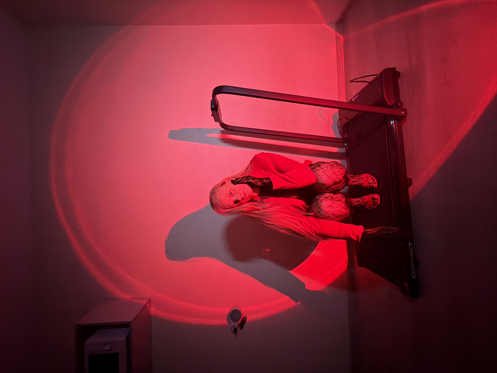 A woman with long blonde hair is squatting on a black treadmill in a room illuminated by red light. The red light casts a large circular glow on the wall behind her, as well as a shadow of her body. She is wearing a red jacket, patterned tights, and open-toed shoes. On the wall behind her, to the left, is a white microwave. The treadmill has the word "COMPACT" printed on its side. The floor appears to be a light-colored wood. The environment suggests an indoor, possibly residential setting in Izhevsk, Russia.
