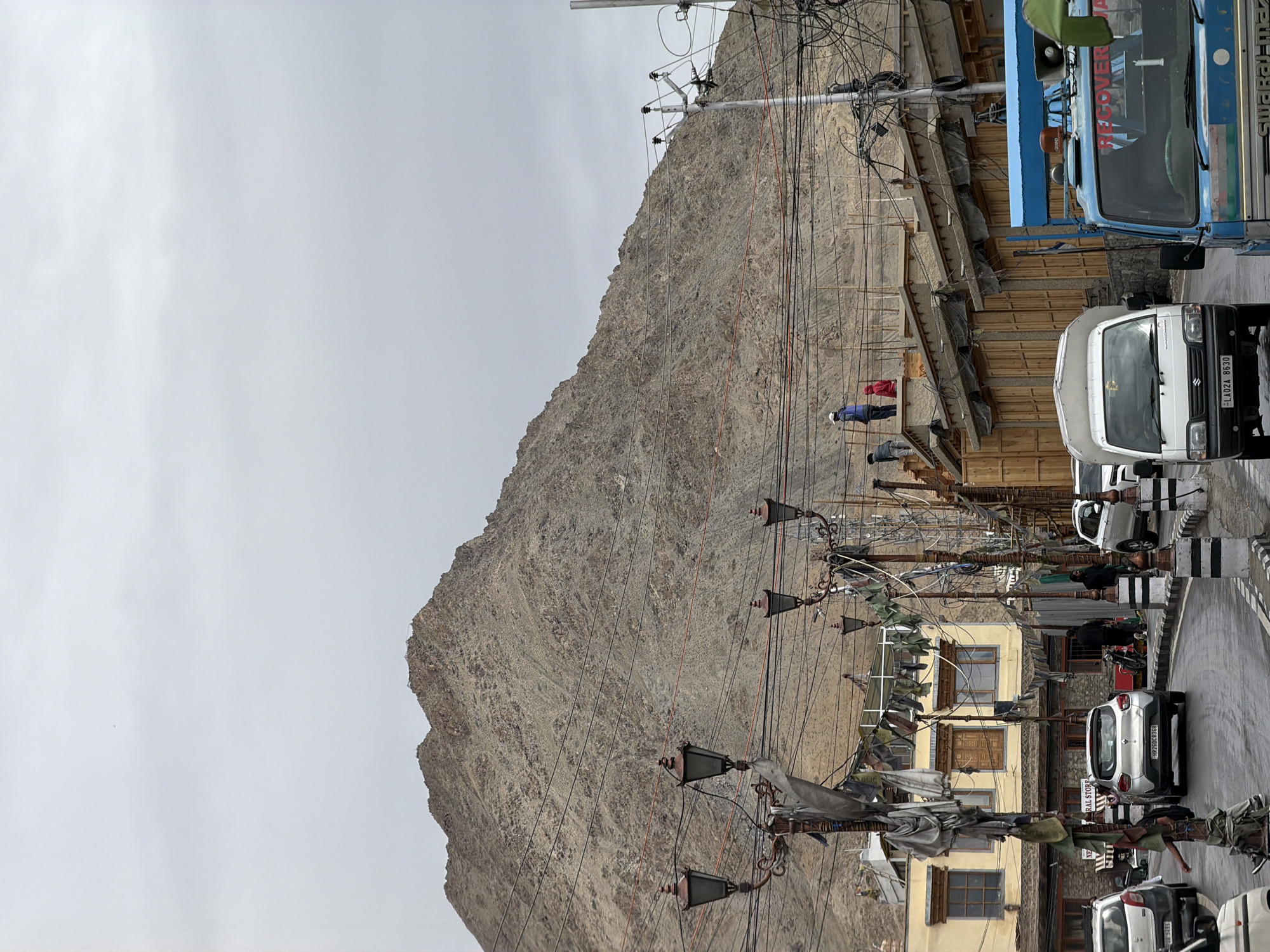 The image captures a street scene in Leh, India, under a cloudy sky. A large, barren mountain dominates the background, its rocky slopes textured with erosion. In the foreground, a paved road curves through the town. Several vehicles, including cars, a white van, and a blue truck, are visible on the road. Buildings with traditional architecture, featuring wooden accents and small windows, line the street. Decorative street lamps with multiple arms and ornate designs are placed along the roadside, adorned with prayer flags or similar festive decorations. Power lines crisscross the scene, contributing to a sense of urban infrastructure against the natural mountain backdrop. Several individuals are present, some seemingly working on the rooftops of buildings under construction, while others are walking or standing near the vehicles. The overall impression is of a busy street in a mountainous region.