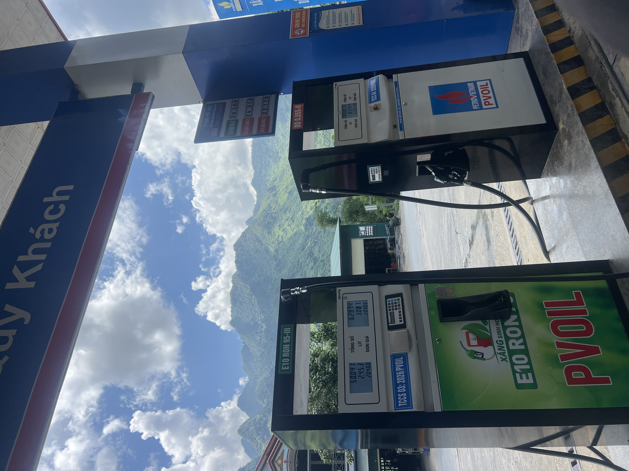 The image shows a gas station in Lùng Tám Commune, Vietnam, with a scenic mountainous backdrop. Two PVOIL gas pumps are visible, one on the left with a green and white design featuring "E10 RON" branding, and another on the right with a white and blue design and the "PETROVIETNAM PVOIL" logo. Both pumps have digital displays showing fuel prices and quantities. The gas station canopy is blue and white, with a large sign at the top partially visible that reads "Quý Khách" (meaning "Guest" or "Customer" in Vietnamese). In the background, lush green mountains rise against a sky filled with white clouds. A building with a green roof and garages is situated behind the pumps. The ground is paved, with yellow and black striped markings near the edge of the platform. No people are clearly visible in the frame.