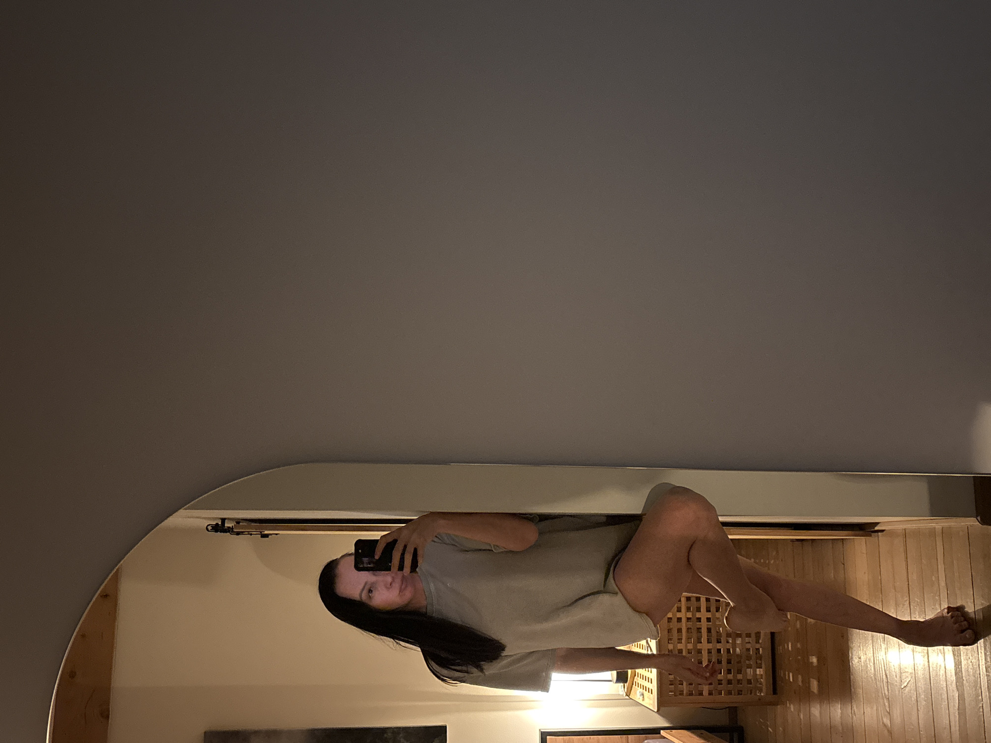 A reflected image in a mirror shows a woman taking a selfie. The woman has long, dark hair and is wearing a light-colored t-shirt. She is holding a phone in her right hand, capturing her reflection. The reflection shows a room with wood floors, a wooden structure, and some artwork on the wall. The mirror is arched, with a metallic track visible above the reflected scene. The setting does not appear to be in Chaikovsky, Russia based on the available information.
