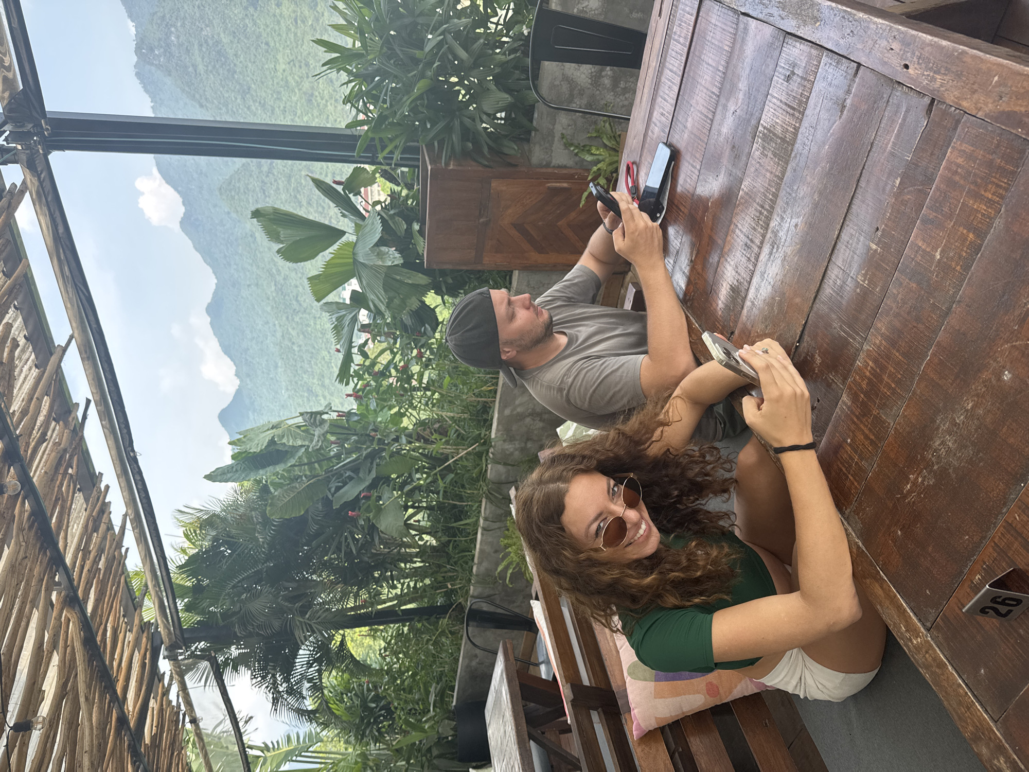 This image captures a vibrant, sunlit outdoor scene featuring two individuals seated at a rustic wooden table against a stunning natural backdrop, indicative of Hà Giang 1 Ward, Vietnam. The setting appears to be a cafe or restaurant terrace, offering panoramic views of lush green mountains under a bright, cloudy sky.
In the foreground, a woman with voluminous curly brown hair is seated, wearing a dark green top, white shorts, and stylish round sunglasses. She holds a smartphone and looks towards the viewer with a broad, cheerful smile, conveying a sense of happiness and relaxation. A black hair tie is visible on her left wrist. Beside her, a man with a beard, clad in a grey t-shirt and a dark cap, is absorbed in his smartphone, looking down intently at the screen. Another smartphone rests on the table near his hands.
The table itself is made of heavy, dark wood with a visible grain and textured surface, suggesting a well-used and natural aesthetic. A colorful cushion with an abstract geometric pattern is placed on the woman's seat. The surroundings are abundant with tropical foliage, including large palm leaves and other broadleaf plants, some adorned with small red flowers, creating a verdant environment. To the left, a vertical structure composed of rough wooden slats, interspersed with string lights, frames the scene, while dark metal beams form part of the overhead canopy. In the distance, layers of majestic green mountains rise under a bright sky dotted with soft white clouds, emphasizing the area's picturesque geography. A small metal tag with the number "26" is affixed to the side of the table, likely indicating a table number. The overall mood is tranquil and leisurely, highlighting a moment of connection with both technology and nature.