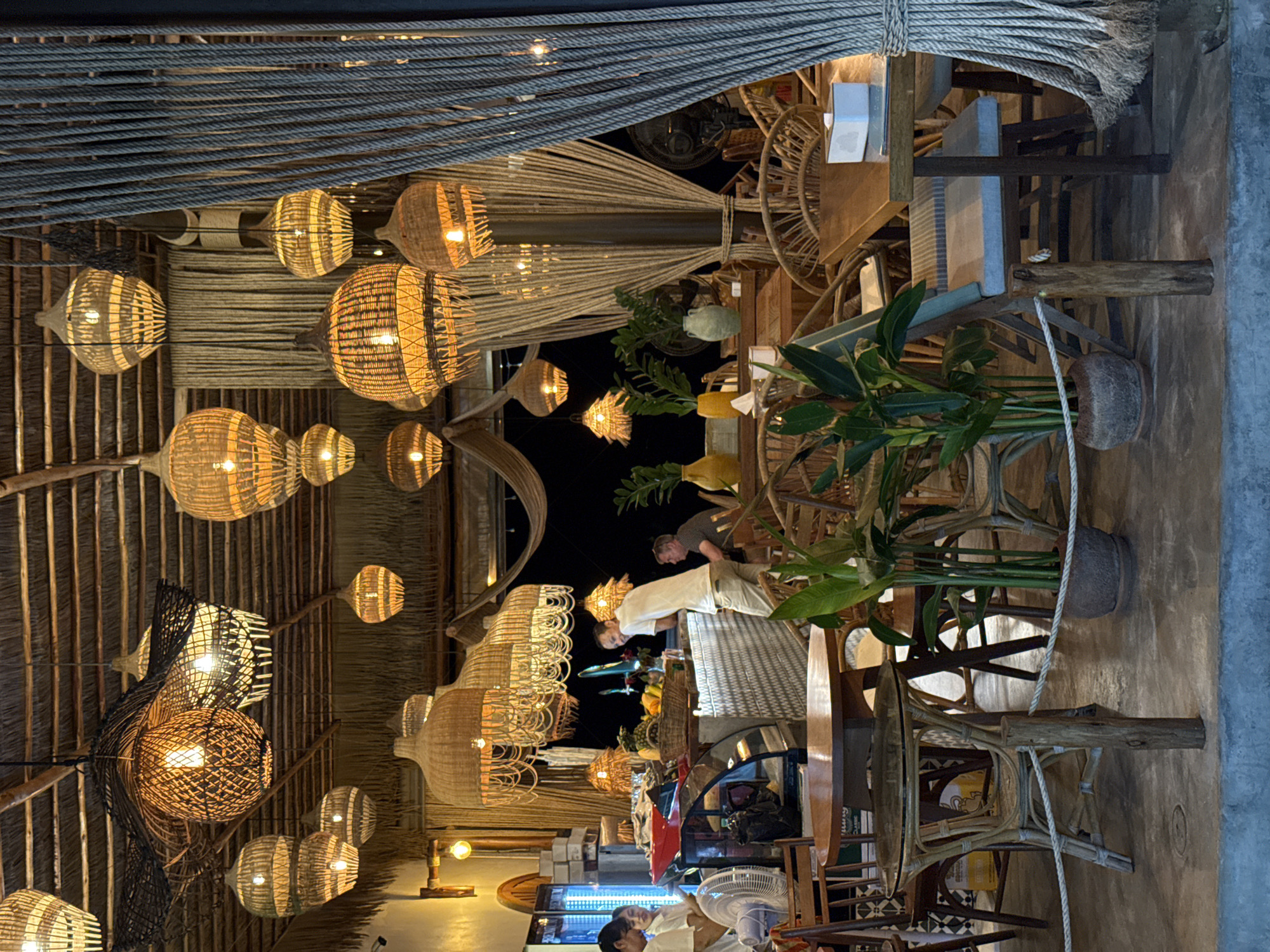 The image depicts the interior of a restaurant in Baan Bo Phut, Thailand, likely a beachfront establishment given the natural materials and tropical ambiance. The ceiling is constructed from woven thatch, with numerous pendant lamps made from rattan and bamboo hanging at various heights, casting a warm, ambient glow. These lamps are a dominant visual element, contributing to the rustic and bohemian atmosphere.
The restaurant features a counter area where two individuals, possibly staff, are visible behind a display case. One, a woman wearing a white top, is partially obscured. Another person, a man in a white t-shirt and light-colored pants, is leaning on the counter, interacting with someone out of frame. To the right, another man is seated at a table, facing away from the camera.
The seating area consists of numerous tables and chairs, predominantly made of rattan and wood, with woven details. Tropical plants in terracotta pots are strategically placed throughout the space, adding to the natural aesthetic. A large roll of thick rope is visible on the right side of the frame, possibly a decorative element or part of the building's structure. Drapes made of thin, woven material are also present, creating dividers and adding texture to the scene. The floor appears to be concrete. The overall impression is one of a relaxed, open-air dining environment, designed to evoke a sense of tropical comfort.
