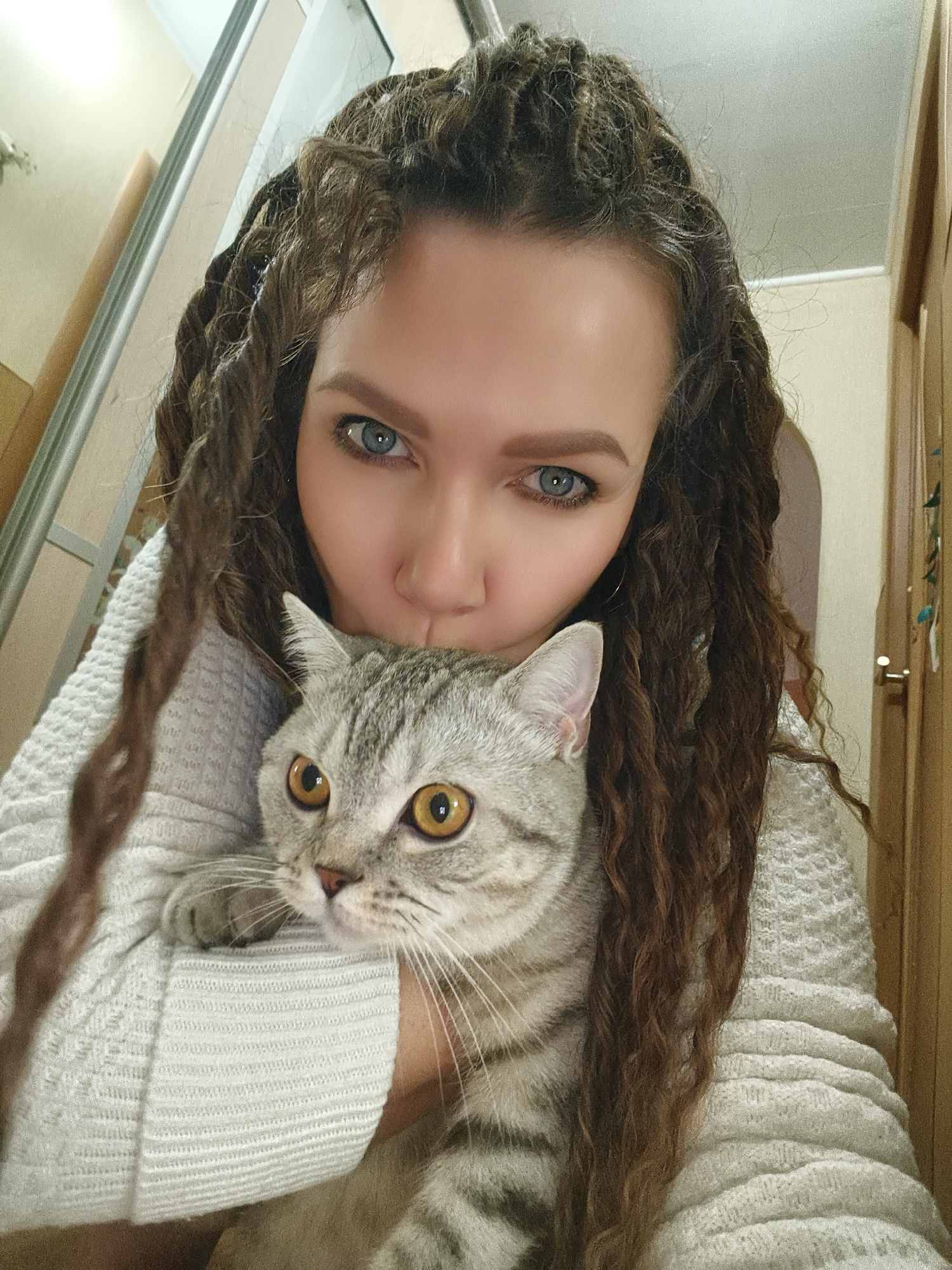 A woman with long, brown braided hair holds a gray tabby cat close to her face. She wears a white sweater and has blue eyes. The background appears to be an interior setting, possibly a hallway with a closet and a door. The location is Unknown, Unknown.