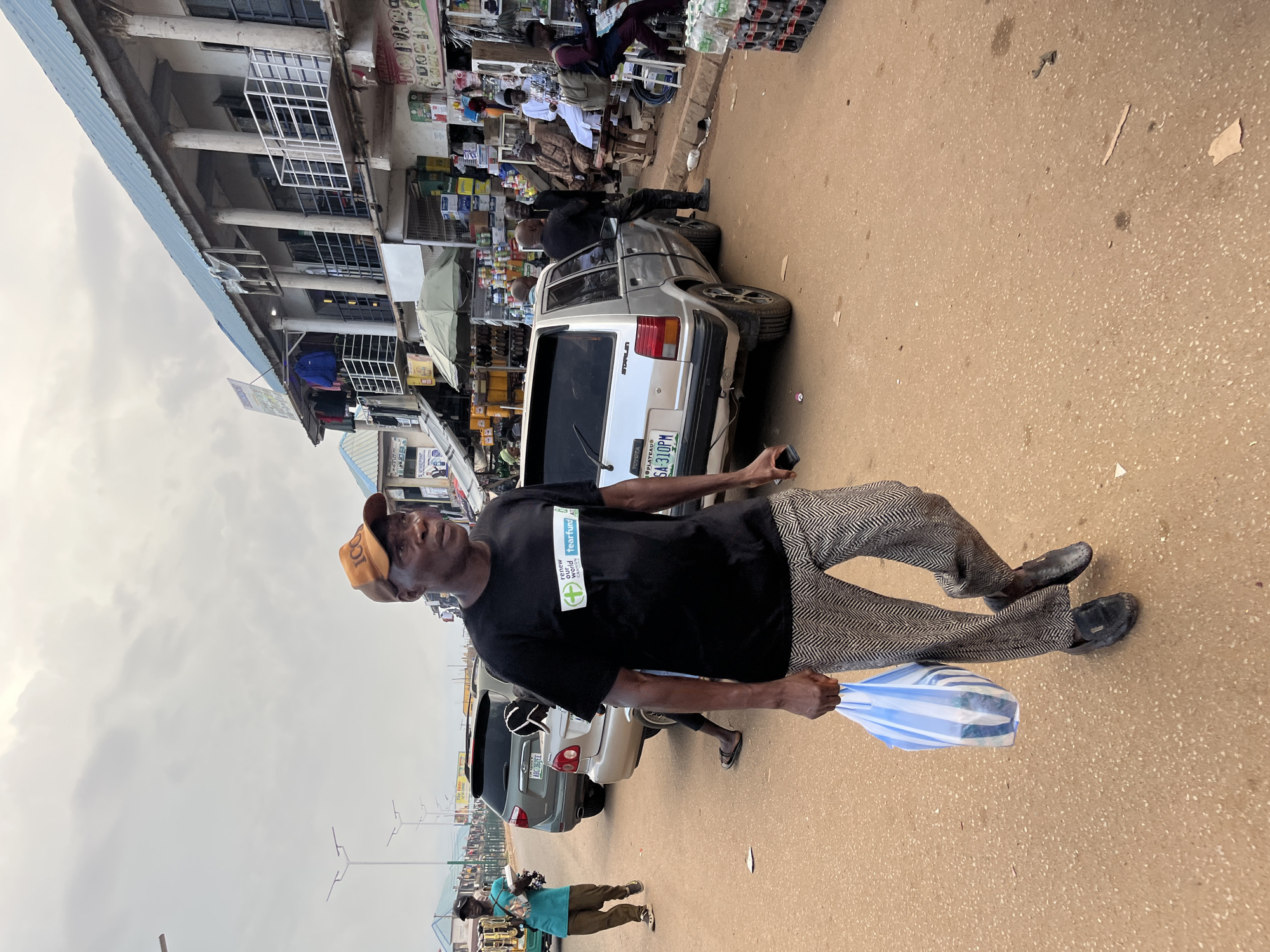 The image depicts a street scene in Bukuru, Nigeria, with a man in the foreground walking and carrying a striped bag. He is wearing a black t-shirt with a logo, a baseball cap, and patterned trousers. The shirt has the text "Tearfund" and "renew our world, change lives" visible. The background shows a busy street with buildings lining one side and parked cars, including a white car with a license plate that reads "SA 310PM". Other people are visible in the background, some near stalls or shops, suggesting a market or commercial area. The weather appears overcast, and the time of day seems to be daytime. The overall impression is a glimpse into daily life in a Nigerian town.