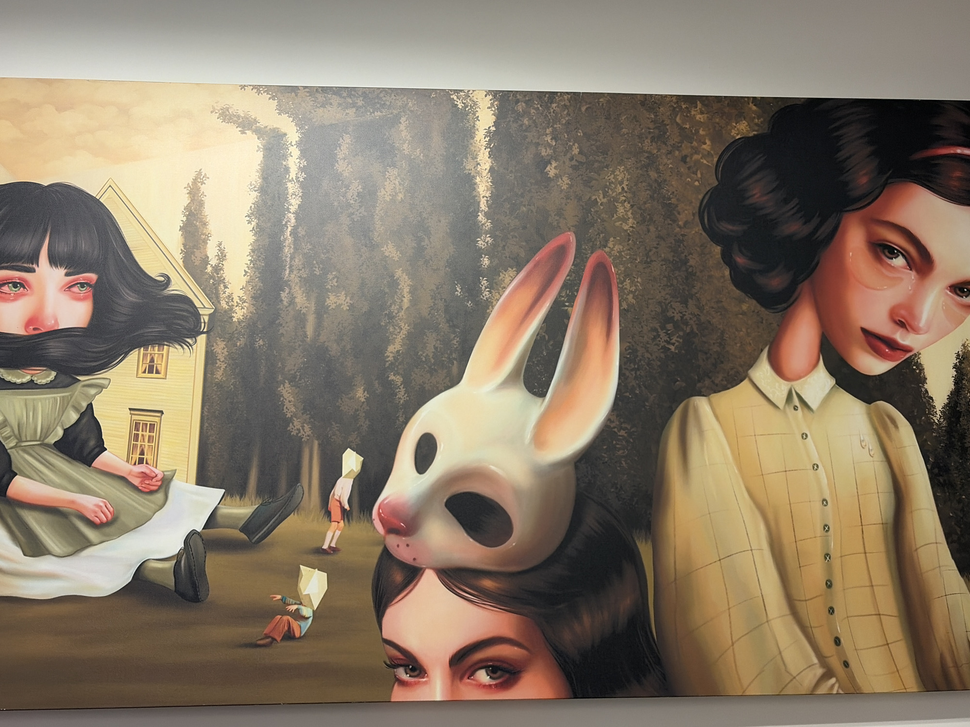 The image shows a large painting.
The painting contains three female figures. On the left, a woman with black hair is sitting on the ground near a yellow house. The bottom half of her face is obscured by her hair, but her eyes look tired and red. Two children with paper bags on their heads are in the background.
In the center, another woman wears a large rabbit mask on her head, with only her eyes visible.
On the right, a woman with dark, styled hair and a yellow checkered dress looks downward with a sad expression.
Behind the figures is a densely wooded area.
The painting is likely in a gallery, but the image doesn't reveal its location in Izhevsk.