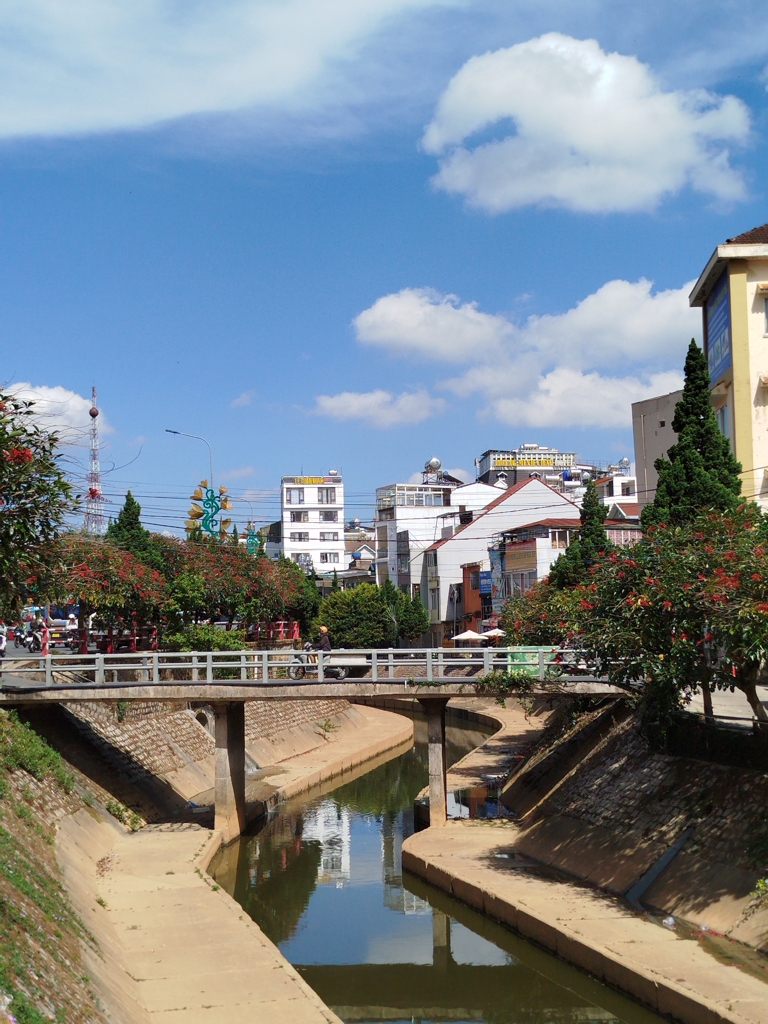 A canal bisects the foreground of an urban scene in Da Lat, Vietnam. The canal is lined with concrete, and spanned by a concrete bridge with a white railing. A few people and mopeds traverse the bridge. Buildings of varying heights and architectural styles comprise the background, with a communication tower visible to the left. The sky is blue with scattered white clouds. Trees with red flowers and green foliage line the bridge, adding a touch of color to the scene. The canal water reflects the buildings and sky.