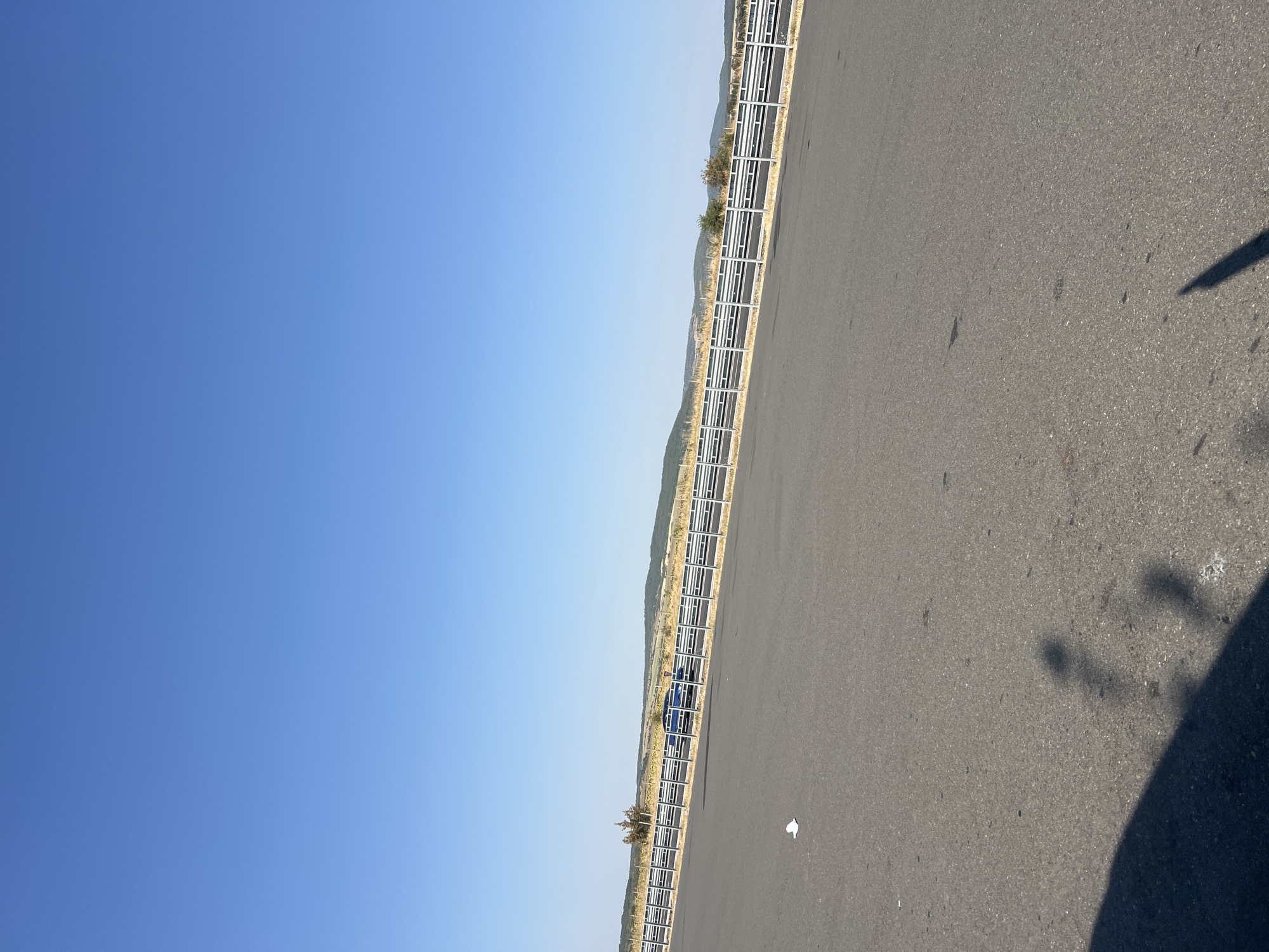 An asphalt road runs diagonally across the frame under a clear blue sky. A metallic guardrail with horizontal bars separates the road from a grassy embankment with scattered trees. In the distance, rolling hills are visible. A blue car is parked on the other side of the guardrail. A white piece of paper lies on the asphalt in the foreground, and shadows of people are cast on the road. The scene appears to be in Vinarovo, Bulgaria.