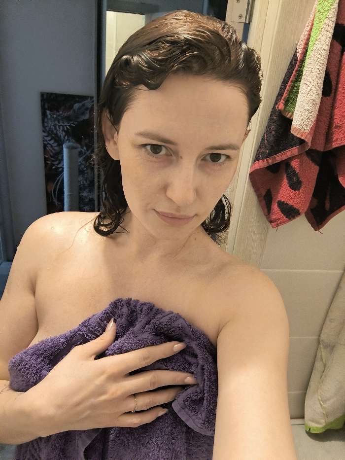 A single adult female, with wet, dark, wavy hair and bare shoulders, is depicted in what appears to be a bathroom. She holds a purple terry cloth towel across her chest with her left hand, which features a thin gold-colored ring on the ring finger and light-colored manicured fingernails. Her gaze is directed at the camera with a neutral expression. To the left of the subject, a dark-framed rectangular object, possibly a mirror or artwork, is partially visible against a light wall, along with a vertical, cylindrical object below it. To the right, multiple towels, including one with a red background and black polka-dot pattern, hang on a light-colored door or cabinet panel. The hardware of the panel, possibly a hinge, is visible at the top right. The specific location within Saint Petersburg, Russia, is not identifiable from the provided visual information.