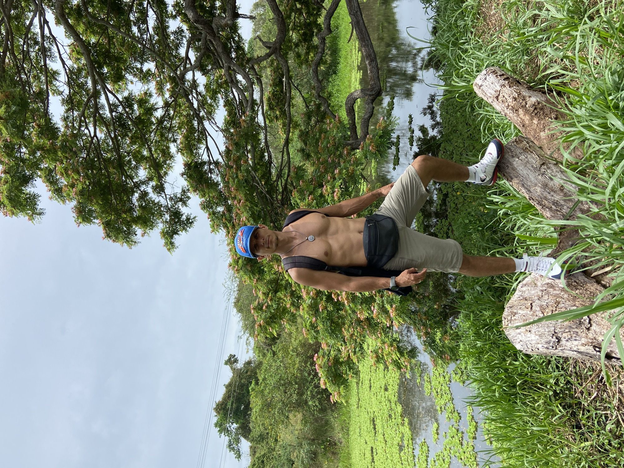 A man wearing a blue cap, necklace, black sling bag, khaki shorts, white socks, and athletic shoes is standing on a log. He is shirtless. The log is situated at the edge of a body of water covered with aquatic vegetation and tall grasses. The scene also includes trees and other lush vegetation. There are power lines in the sky. The location is Río Chico, Venezuela.