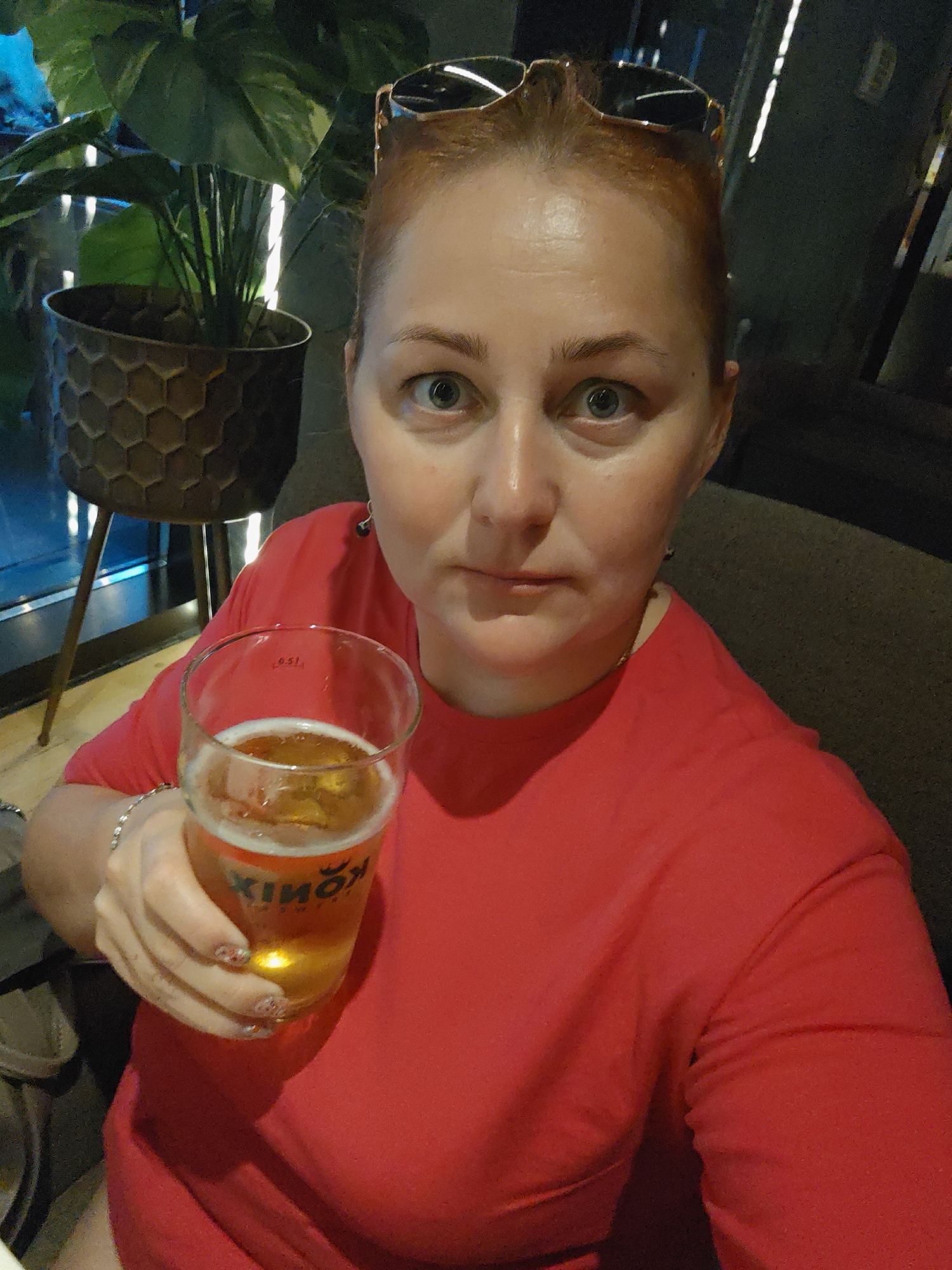 The image depicts a close-up selfie of a fair-skinned woman with reddish-blonde hair pulled back, wearing a bright red t-shirt. She is holding a glass of amber-colored beer in her right hand, with her left arm resting on her lap. Sunglasses are perched on top of her head. Her facial expression is one of mild surprise, with wide eyes. In the background, a potted plant with large green leaves is visible to the left, and dark, blurred surroundings suggest an indoor setting, possibly a bar or restaurant. The logo on the beer glass is partially obscured but appears to be in Cyrillic. The overall impression is of a casual moment captured in a relaxed social environment. Given the Cyrillic script on the beer glass, the image was likely taken in a location where Russian is spoken, such as Saint Petersburg, Russia, possibly in a bar or cafe.