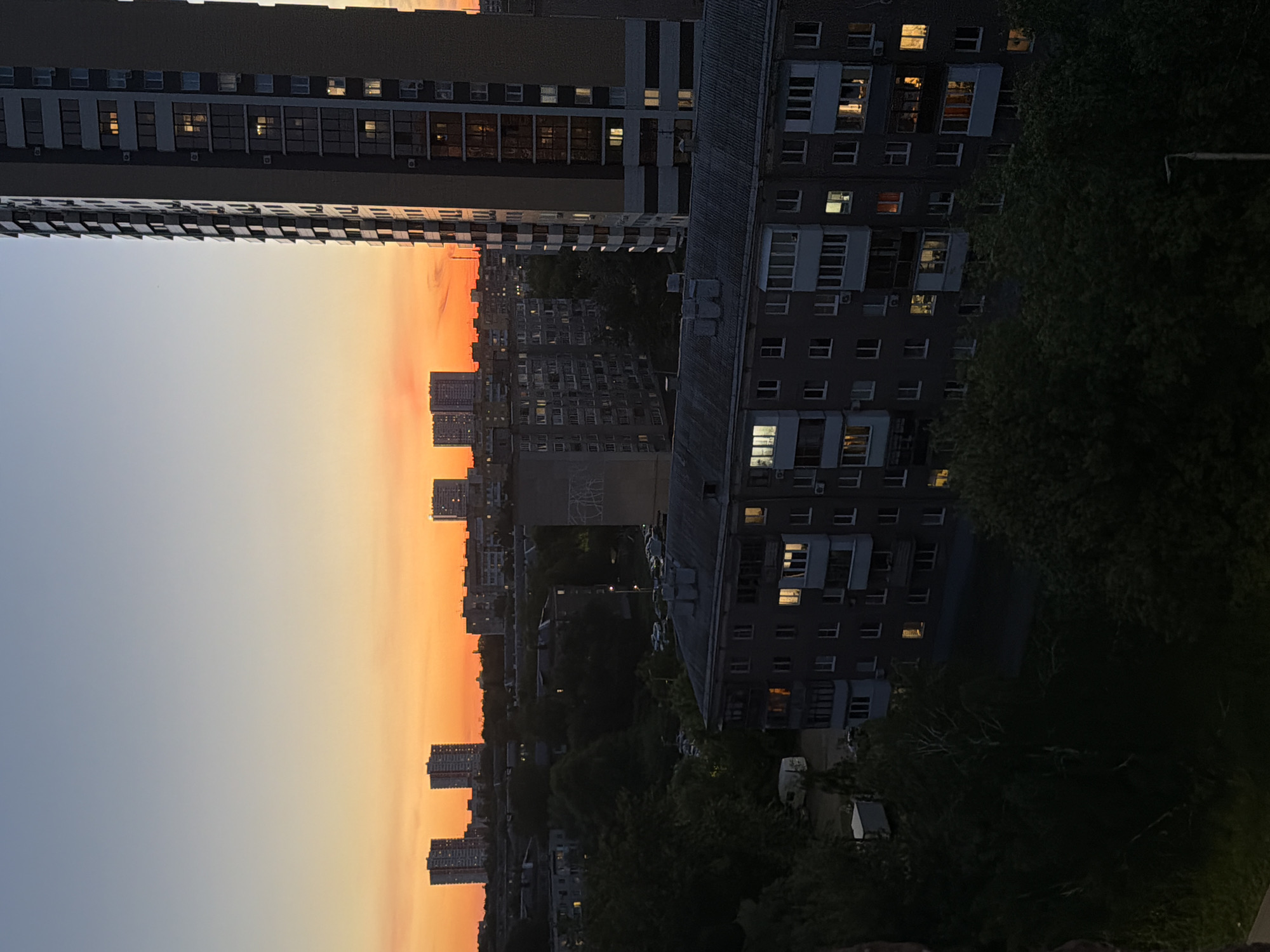 The image shows a cityscape at dusk. In the foreground are apartment buildings, with some windows lit up. Between the buildings is a view of other high-rise buildings in the distance, silhouetted against an orange and blue sky. There are also trees visible.