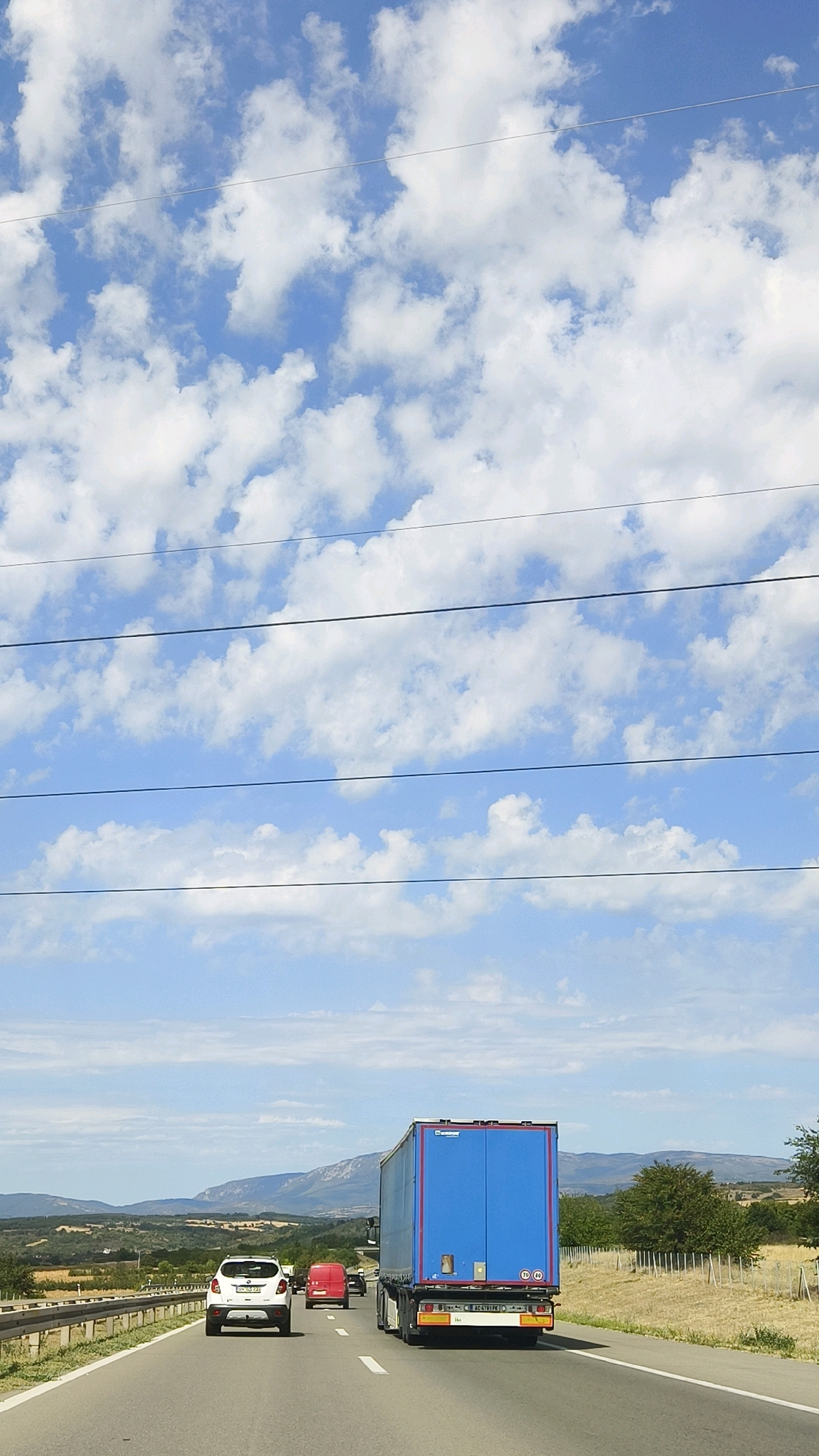 The image shows a road with several vehicles, including a white SUV, a red van, and a blue semi-trailer truck, driving on a highway. The scene is set against a backdrop of a blue sky with white clouds and rolling hills in the distance, indicating an outdoor environment likely in a rural or suburban area. The presence of utility lines overhead suggests the road is a main thoroughfare. Based on the visual cues and the provided location, the image depicts a section of a highway within or near the municipality of Crveni Krst in Serbia, where vehicular traffic is observed moving along the paved road under a cloudy sky.