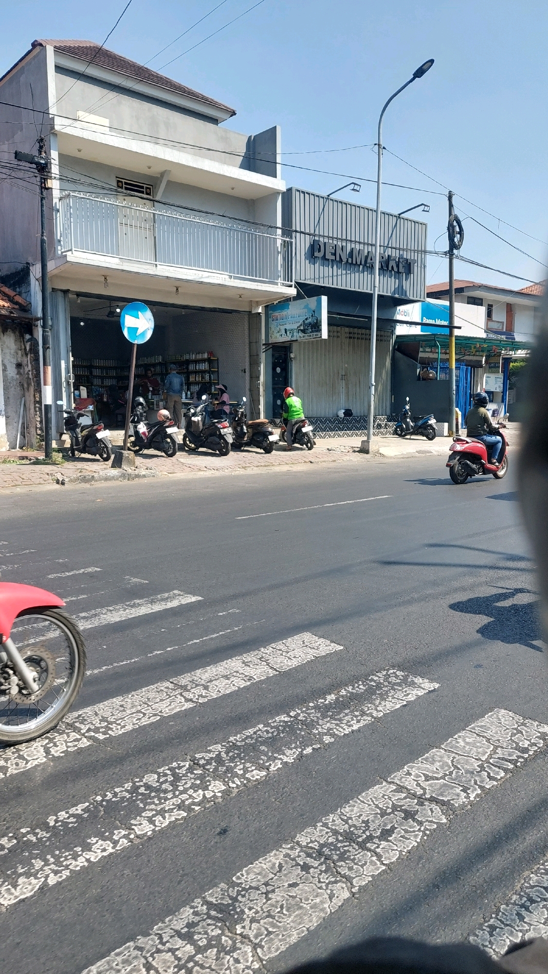 The image depicts a street scene in Sidoarjo, Indonesia, on a sunny day. A crosswalk is prominently featured in the foreground, with a motorcycle wheel partially visible on the left side. In the background, a two-story building with a white balcony is visible, adjacent to a building with a metal facade bearing the text "DEN MARKET". Several motorcycles are parked in front of the buildings. People are seen near the entrance of the "DEN MARKET" building. One person in a green jacket is riding a red scooter across the street, and another person in a brown jacket is riding a scooter. A blue directional sign with a white arrow is also visible on the sidewalk. The environment suggests a commercial area with moderate traffic and pedestrian activity.