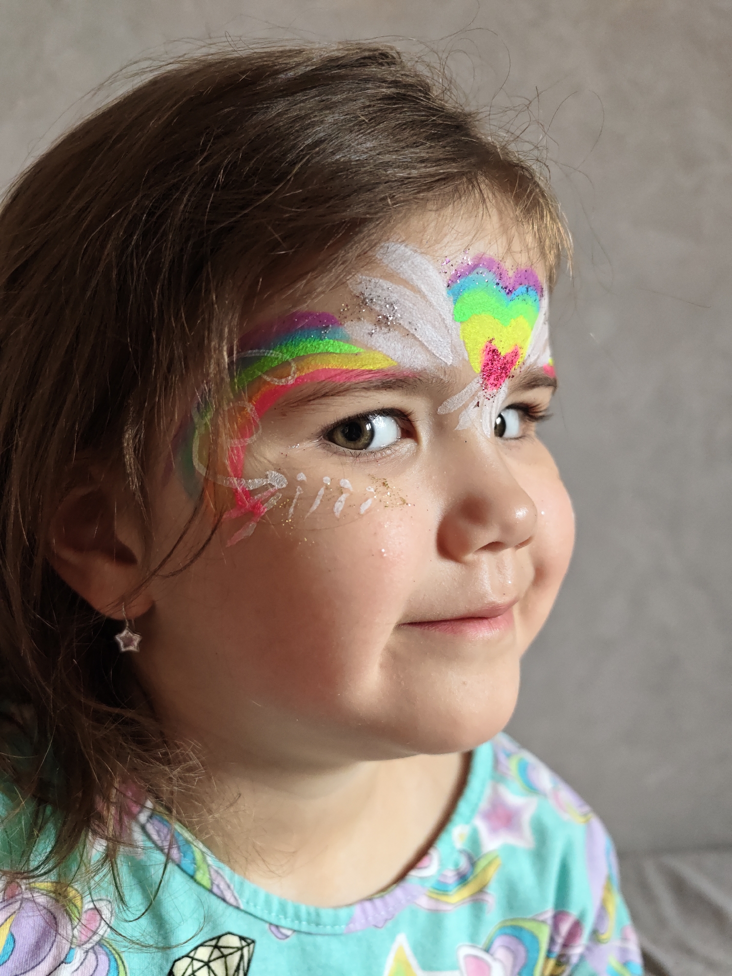 A close-up portrait depicts a young child, likely female, with light brown hair and brown eyes, positioned off-center looking towards the right of the frame with a slight smile. Her face is extensively painted; a rainbow-colored arc, accented with white wing-like patterns and silver glitter, extends across her forehead above her left eye. A small pink heart shape is centered at the top of her nose bridge, beneath the rainbow. Additional white dots and lines, alongside gold glitter, are applied to her cheeks below both eyes. A small, silver-colored star-shaped earring is visible in her left earlobe. She wears a light teal garment featuring colorful patterns of stars, unicorns, and geometric shapes. The background is a uniformly textured, out-of-focus light grey surface. There are no visual elements within the image to specify a location within the city of Chaikovsky, Russia.