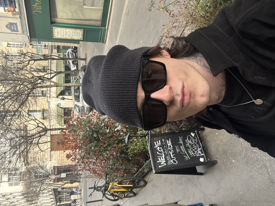 The image presents a male individual taking a selfie on a sidewalk in Paris, France, specifically near Rue Amelot.
The subject is a male with dark wavy hair, wearing a black knit beanie, black square-framed sunglasses, a black collared jacket, and a black t-shirt. A silver chain with a circular pendant is visible around his neck. Tattoos are present on his neck. His lips are slightly puckered.
To the subject's left, a black A-frame chalkboard sign is positioned on the sidewalk. It displays white chalk writing that reads "WELCOME !!! PLEASE WAIT IN LINE OUTSIDE" at the top, followed by "TODAY: HOT GINGER LEMON HONEY DRINK" and three smiling cartoon faces at the bottom.
In the midground, a rectangular planter box containing green and reddish-brown foliage separates the sidewalk from the street. Two bicycles are parked adjacent to the planter, one yellow and one dark-colored.
The background reveals a typical Parisian street scene with multi-story, light-colored Haussmannian buildings. Bare trees line the street. A street sign identifying "RUE AMELOT" is affixed to a building on the right. Commercial premises with green facades and signs indicating "DELICATESSEN CAVE" and "VITRE" are visible. A "PAN BAGNAT" sign is discernible further down the street on the left. Several vehicles, including a silver hatchback and a grey SUV, are parked or in motion. A "30" km/h speed limit sign is visible on a pole. The sky is overcast. Limited pedestrian activity is observed in the distance.