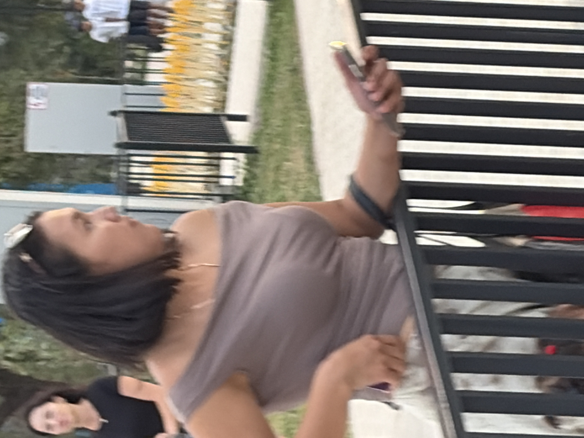 In the foreground, a woman with dark hair and a grey off-the-shoulder top is partially visible, looking to her right and holding a light-colored object that appears to be a phone. Her left arm is bent, and her hand is near her chest. She is standing behind a dark metal fence with vertical bars. Her lower body is obscured by the fence and a patterned skirt or dress. In the background, a woman in a black t-shirt is visible further to the left, and to the right, three young males in white t-shirts and dark shorts are standing in a line. They appear to be in an outdoor public area with a paved walkway and grassy patches, with trees and yellow decorative elements visible behind the fence. There is a light-colored rectangular structure in the background with a red and white sign.