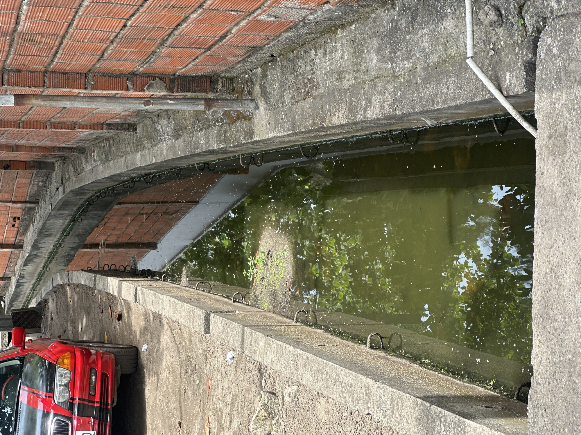 The image depicts a concrete drainage canal filled with green water, reflecting trees. Above the canal is a brick archway. A red van is partially visible on the left side of the frame. The image is rotated counterclockwise. There is no indication that the image depicts Lagos, Nigeria.