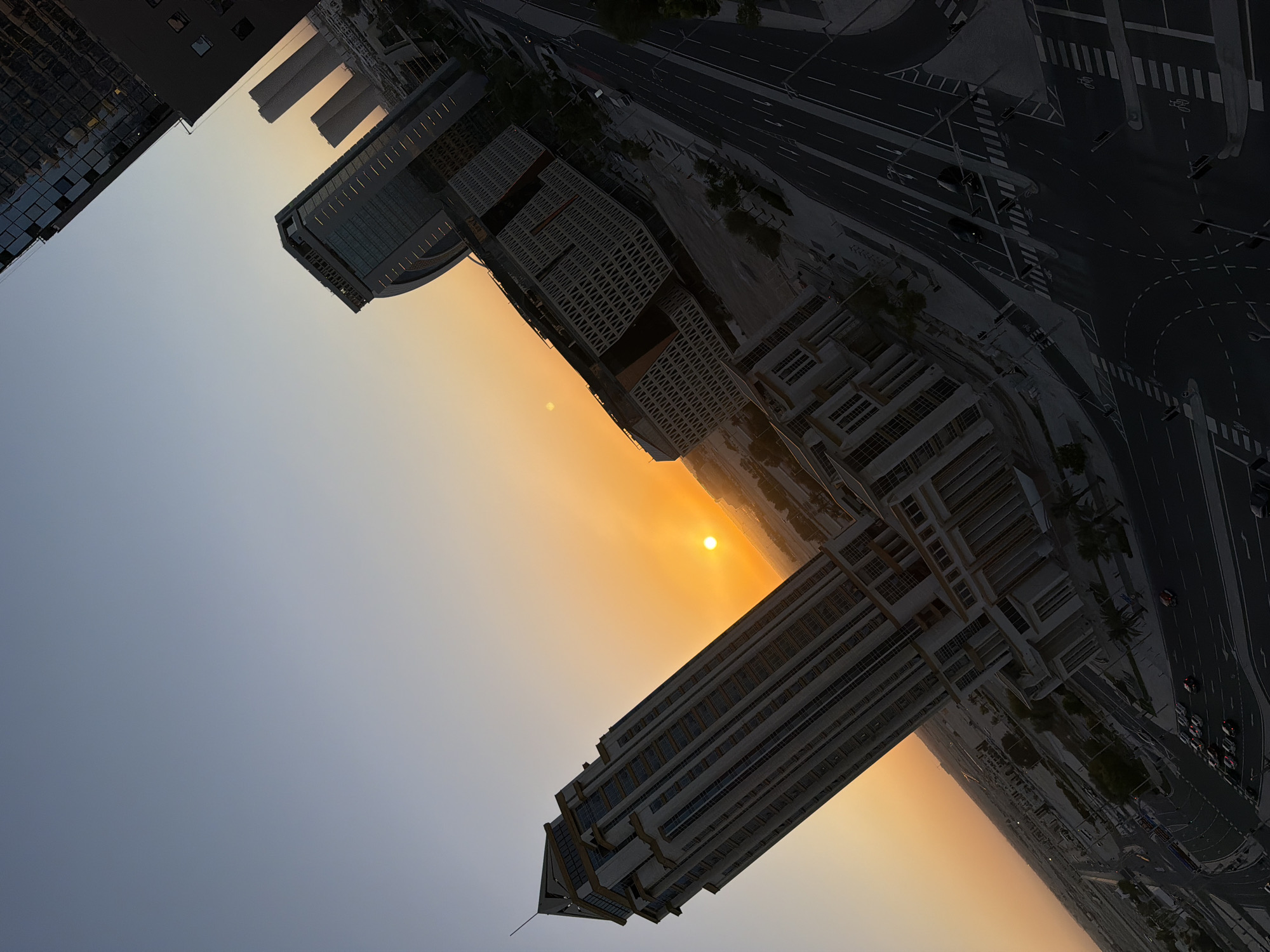 This media file is an image showing a city skyline at sunset. The main subjects are modern skyscrapers and buildings, roads with traffic, and the sky. The sun is visible as a bright orb low on the horizon, casting a warm orange glow across the sky and illuminating the edges of the buildings. The scene is urban, with a clear indication of a developed city.
The activity taking place is the transition from day to night, with the sun setting and lights beginning to appear in the city. Cars are seen driving on the roads, indicating normal city life. There are no visible people.
Notable details include the time of day, which is clearly sunset, as evidenced by the color of the sky and the position of the sun. The weather appears clear, with no clouds obscuring the sunset. The architecture of the buildings suggests a contemporary urban environment. The overall mood conveyed by the image is one of serenity and the beauty of a city at dusk.
There is no visible text in the image.