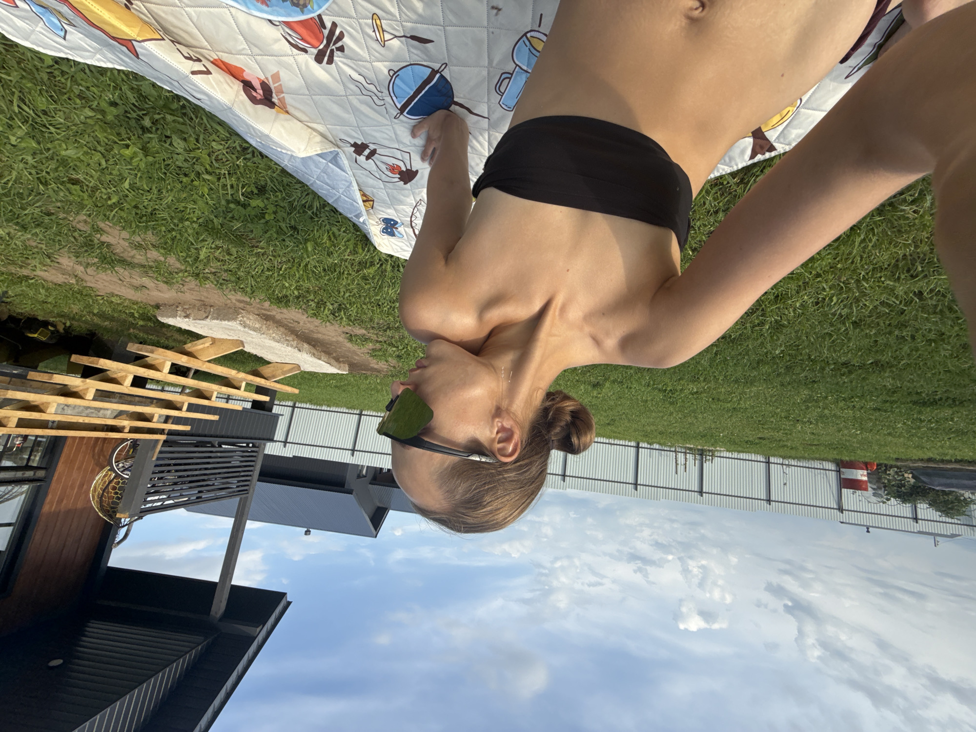 In the city of Podshivalovo, Russia, a young woman with her hair in a bun is sunbathing on a patterned blanket in a grassy yard. She is wearing a black bandeau bikini top and large, dark sunglasses. She is lying on her side, looking away from the camera. In the background, a modern wooden house with a dark roof and a porch is visible, along with a metal fence and lush green grass. The sky is partly cloudy, with a mix of white clouds and blue sky. There are some wooden steps and building materials near the house. The overall scene depicts a relaxed, outdoor setting.