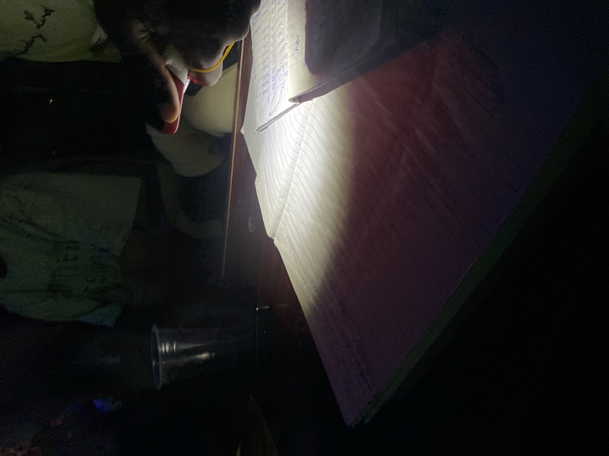 An indoor scene in the city of Howayi, Nigeria, characterized by extreme low-light conditions. In the foreground, a dark-skinned hand is prominently featured, holding a red and white writing utensil and directing a focused beam of white light from a small handheld device (likely a smartphone flashlight) onto an open, lined notebook. The notebook, positioned on a dark wooden table, displays handwritten text in blue ink across its pages, illuminated by the intense light. To the left of the notebook, a clear plastic cup containing a dark liquid is visible. In the deeply shadowed background, several indistinct human figures are partially discernible. One figure wears a light green shirt with faint dark text, and another appears in lighter-colored attire, indicating the presence of multiple individuals within the same dimly lit space, potentially engaged in study or work under challenging lighting circumstances.