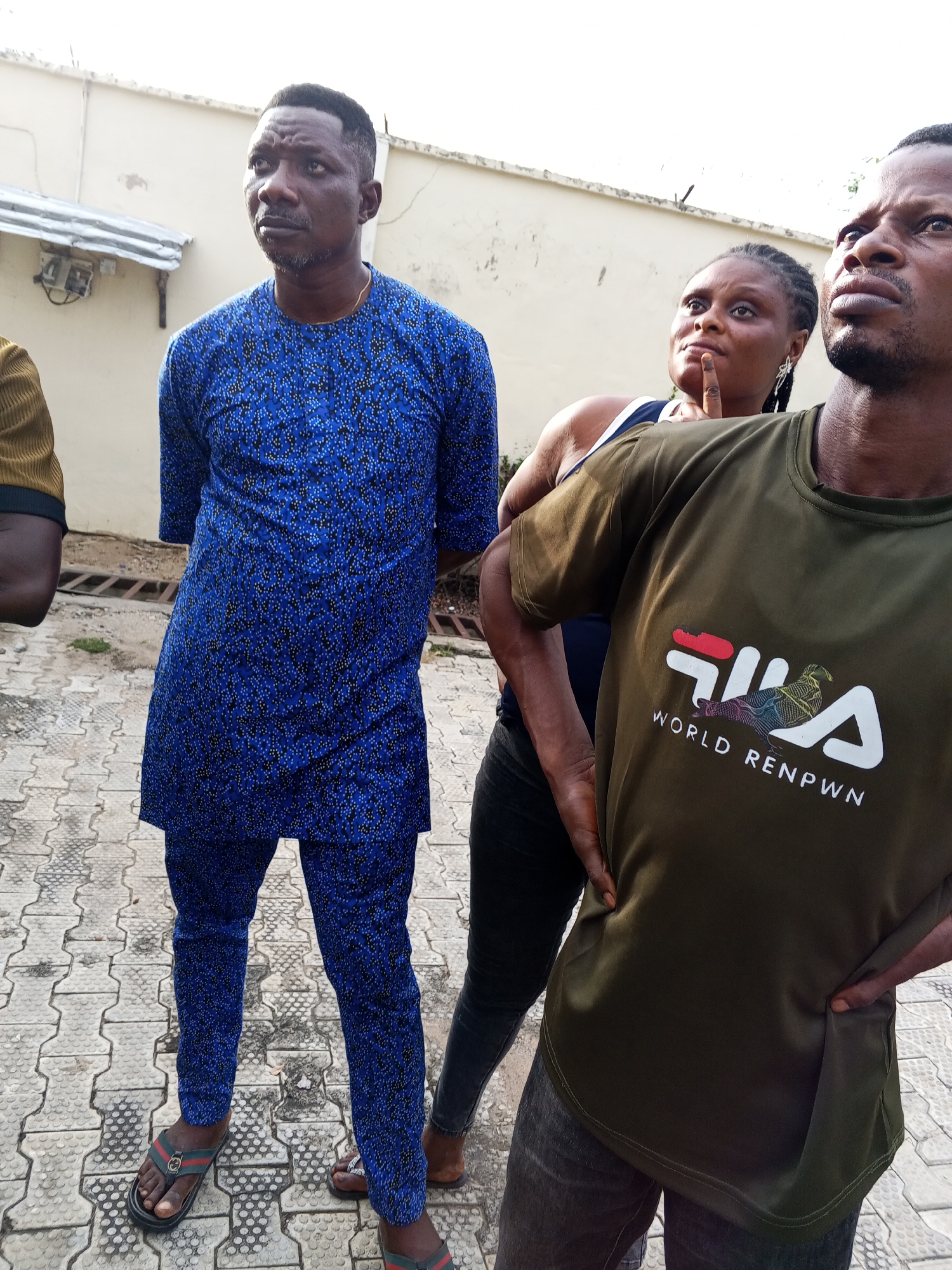 The image shows four people standing outdoors on a paved surface. The person on the left is partially visible and wears a mustard-colored shirt. Next to him is a man in a blue patterned tunic and pants, wearing sandals. A woman stands in the background, appearing to look upwards with a finger raised to her chin. To the right, a man in an olive green "FILA WORLD RENPWN" t-shirt stands with his hands on his hips, also looking upwards.
The setting appears to be a courtyard or similar paved area, with a white wall in the background. Based on the prompt requesting information on the city of Lokoja, Nigeria, and cross-referencing the architectural styles and attire, it is highly probable the location is in Lokoja, Nigeria, although precise identification is not possible without further contextual information. The scene suggests the people are observing or discussing something overhead.