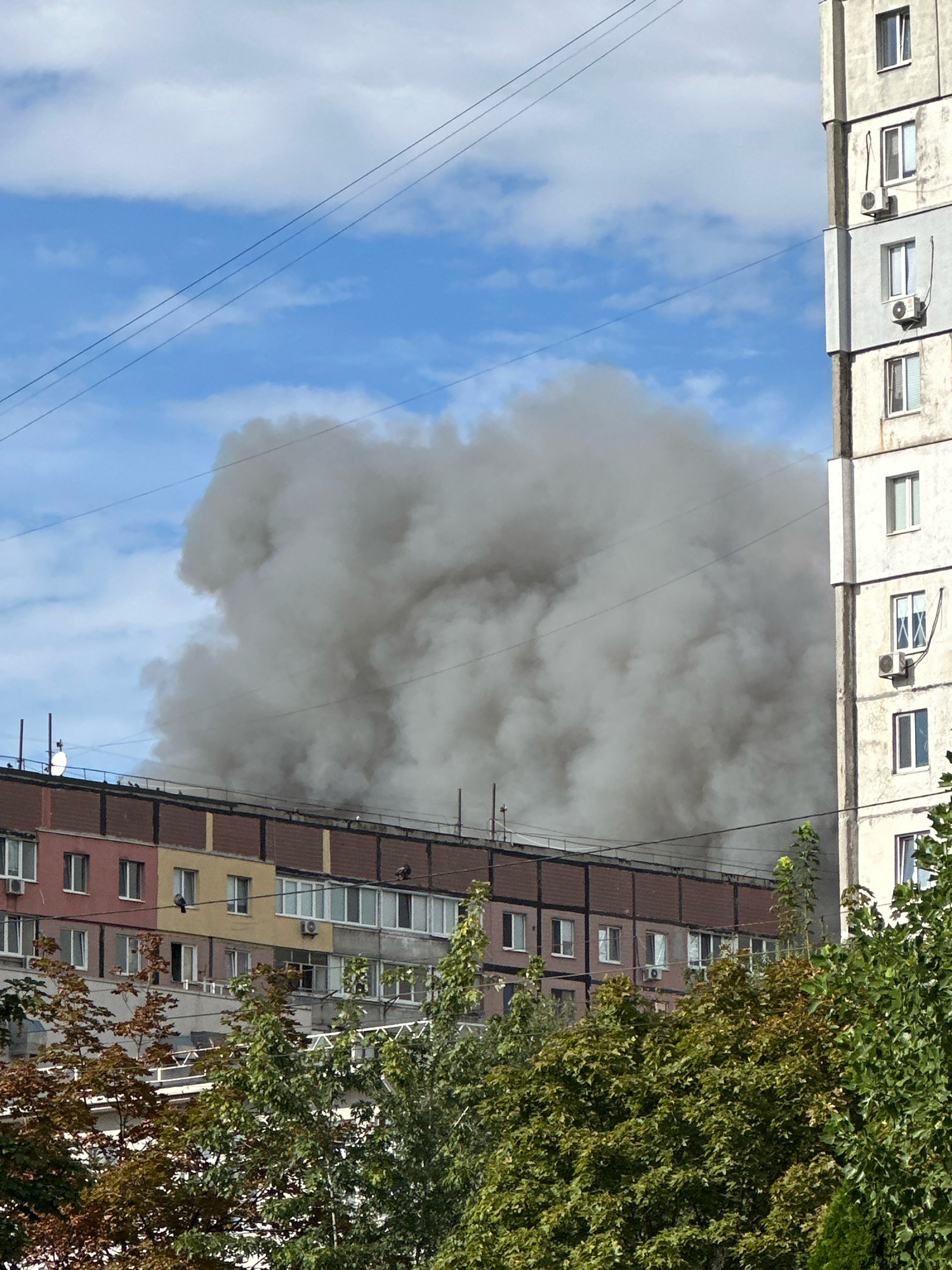 A massive plume of dark gray smoke billows into a bright blue sky filled with scattered white clouds. The smoke originates from behind a long, multi-story apartment building, obscuring a significant portion of its roofline. This building features a facade with sections of red, yellow, and brown paneling, interspersed with numerous windows and balconies. Several air conditioning units are visible mounted on the exterior. To the right of this building, another taller, concrete-faced apartment building is partially visible, also with many windows and air conditioning units. In the foreground, the tops of green and reddish-brown trees can be seen, partially obscuring the lower floors of the apartment buildings. Electrical wires are strung across the sky in the upper portion of the image. The scene appears to be in a residential area of the city of Dnipro, Ukraine, given the context of the event.