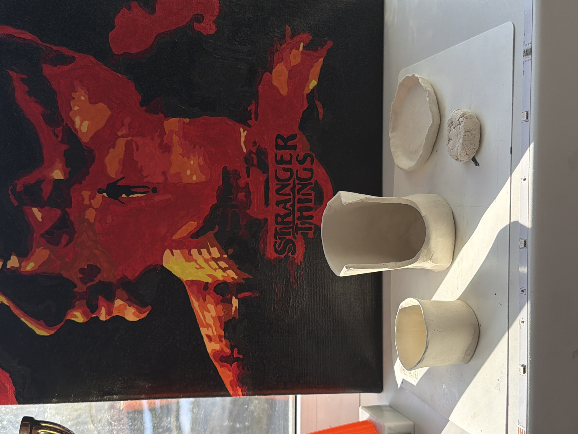 The image shows a close-up of a canvas painting of Vecna from "Stranger Things" next to several small, unfired clay objects on a white surface. The clay objects include a small dish, a textured ball, a larger oval bowl, and a cylinder. The clay objects are illuminated by sunlight. The location in the city of Perm, Russia is not discernible.