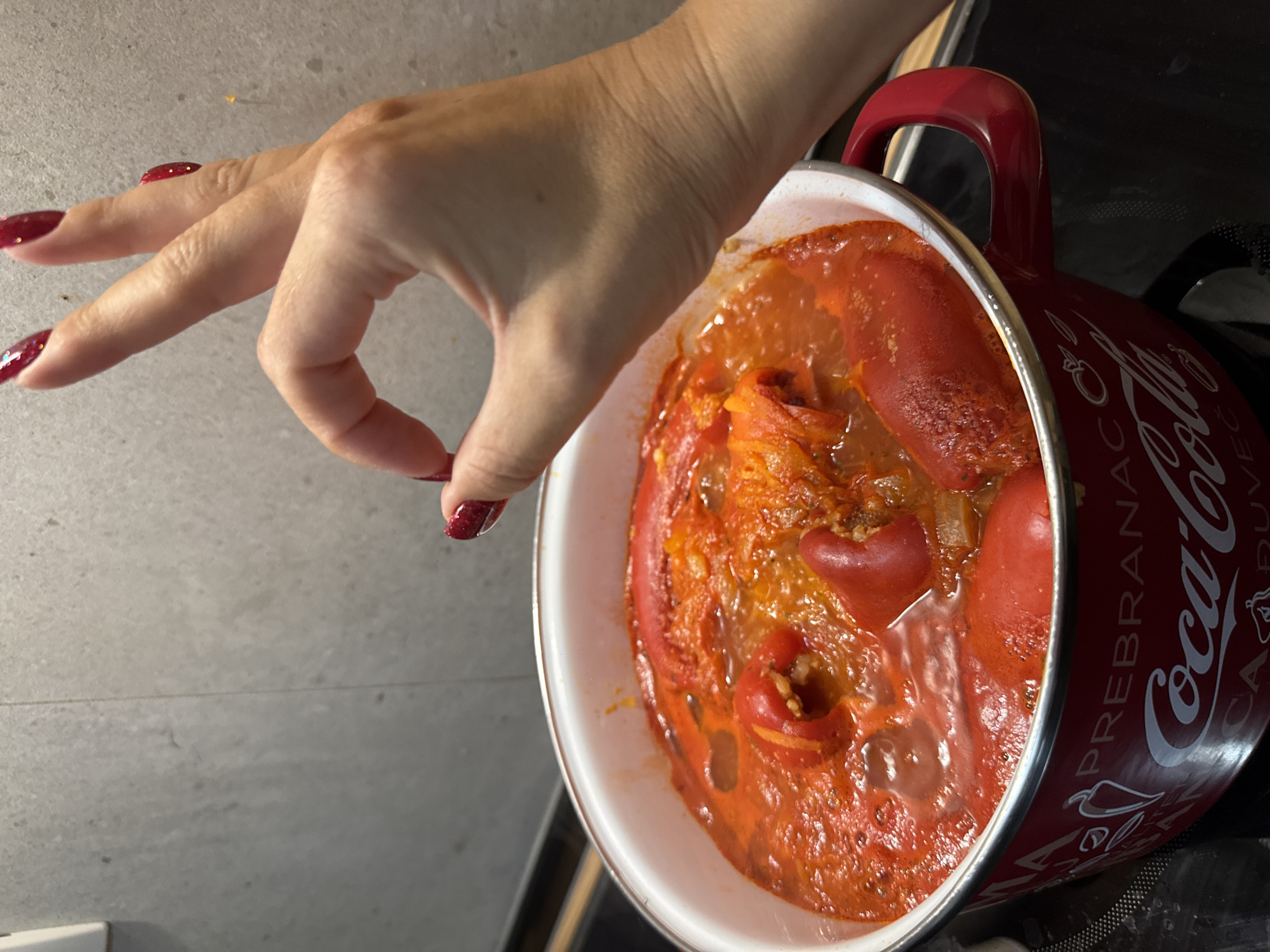 A close-up, eye-level shot captures a red and white enameled pot on a black stovetop, filled with a bubbling, red stew containing pieces of red pepper and possibly meat. A hand with long, red, glittery fingernails enters the frame from the right, forming an "okay" gesture with the thumb and index finger touching. The background is a light gray tiled wall. The pot has "Coca-Cola" branding visible on its side.