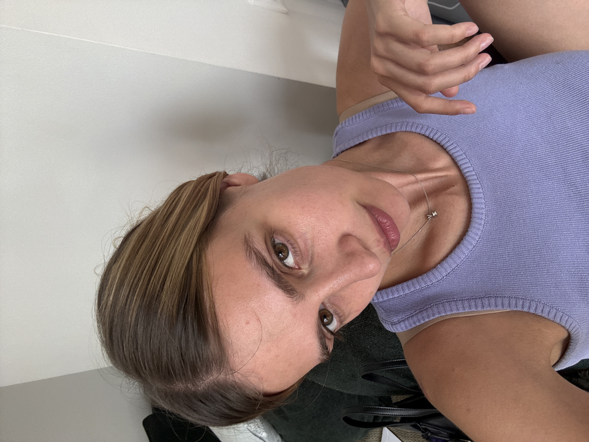 A woman is seen from a close-up, angled perspective. She is wearing a lilac ribbed tank top and a delicate necklace. Her hair is parted to the side and appears to be brown with some lighter highlights. Her skin tone is light, and her eyes are brown. A partially visible hand is positioned above her shoulder. The background includes a plain white surface and portions of other dark-colored objects. The location is in the city of Unknown, Unknown.