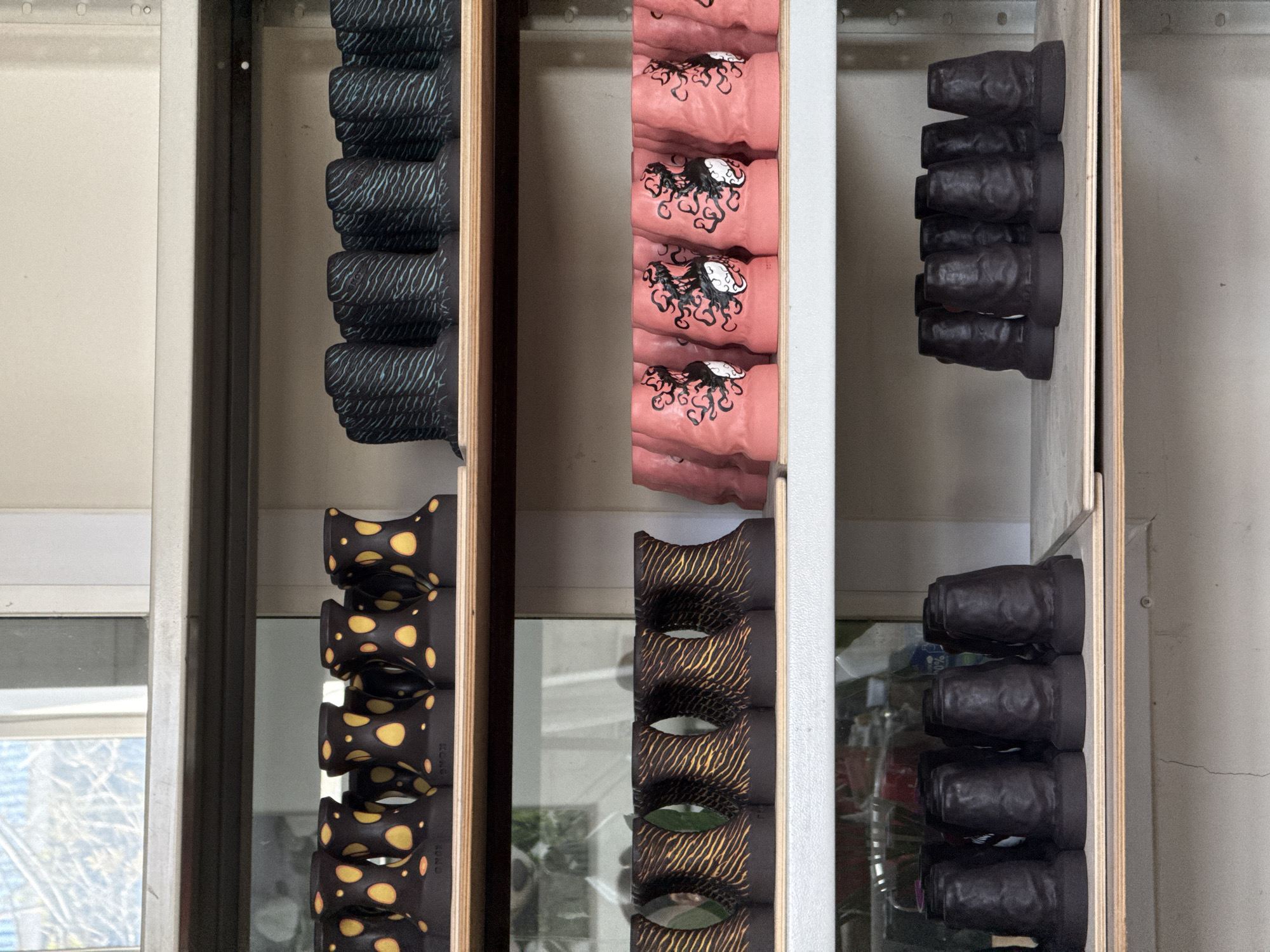 The image shows an open cabinet with three shelves. The leftmost shelf contains vertically stacked objects with a dark pattern on a black or dark-colored surface. The middle shelf contains stacked pink objects with a black octopus design. The rightmost shelf contains stacked dark, matte objects, possibly black or dark grey. The location is Chaikovsky, Russia.