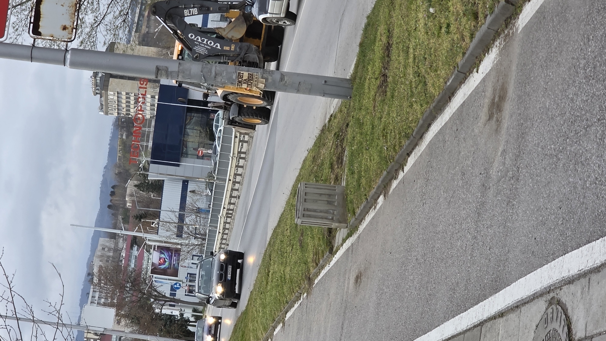 The image shows a street scene in what appears to be Blagoevgrad, Bulgaria, recognizable by the "TECHNOPOLIS" sign on a building in the background. An earthmover branded "VOLVO BL71B" is parked partially on the roadway. There is a car with the license plate "E 8434 HB" moving along the street. Other details include a grey metal object, a grassy strip, trees, a light pole, and utility poles. The sky is overcast. The image appears to be taken from a ground-level perspective.