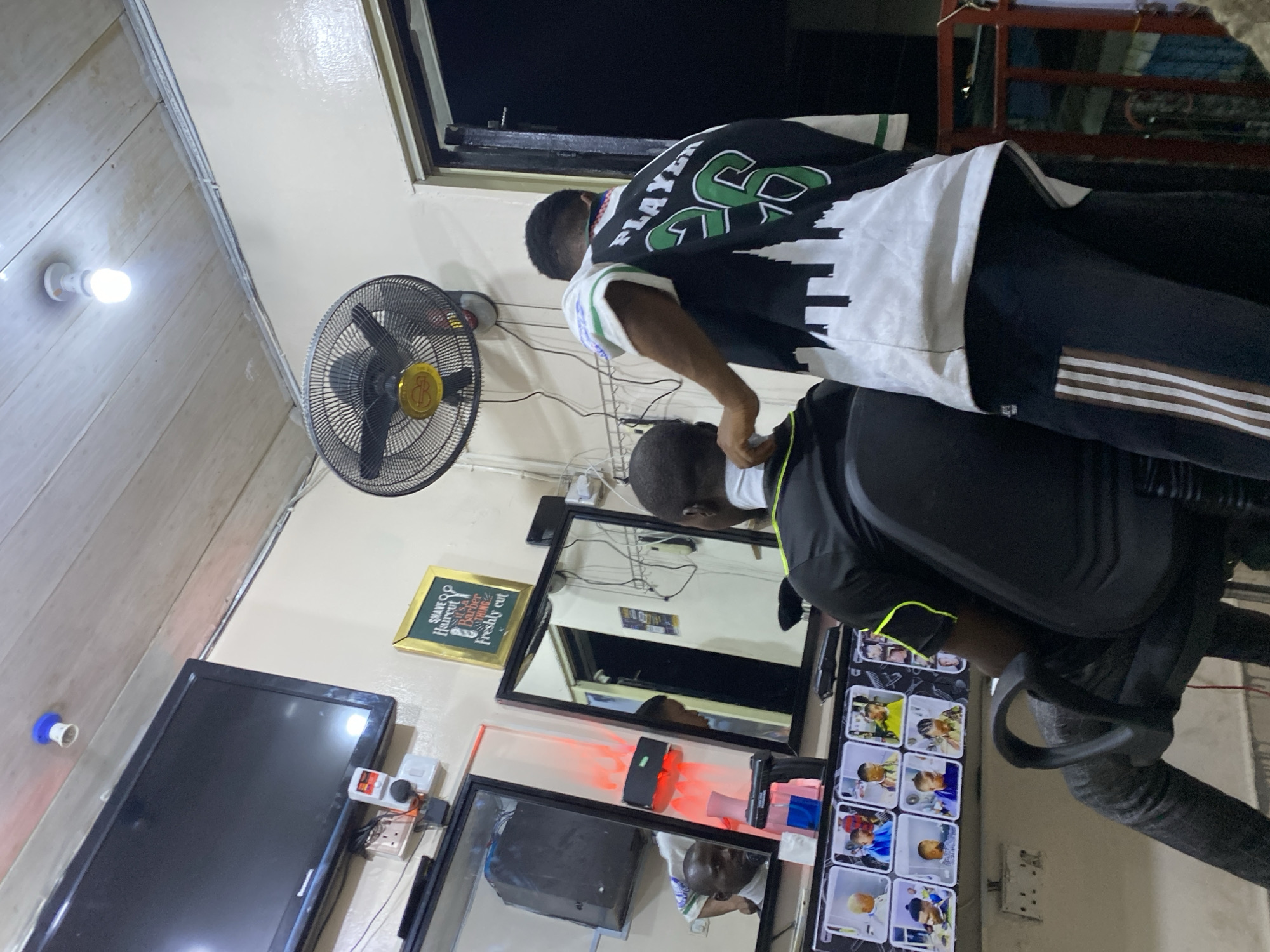 A barber trims a client's hair in a barbershop in Ado, Nigeria. The barber, wearing a green and white sports jersey, stands behind the seated client. The client is wearing a black and white shirt with a yellow outline. Mirrors are mounted on the wall, reflecting the scene. Other items visible include a television, a fan, and an advertisement for haircuts.