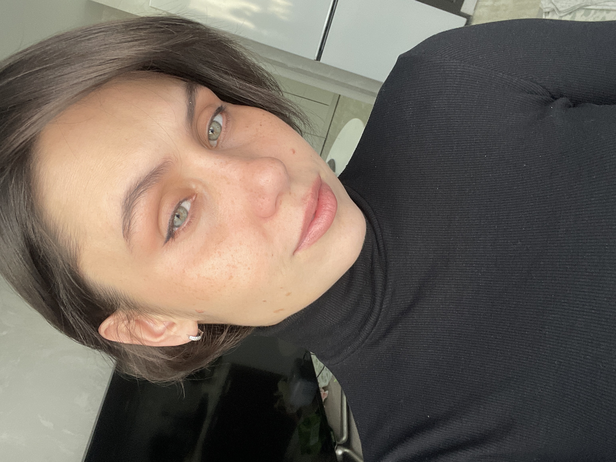 A close-up, angled portrait features a young woman with short, dark brown hair, looking slightly to her right. She has fair skin with numerous freckles across her nose and cheeks, and striking light green eyes. Her eyebrows are well-defined, and her lips are naturally pink and slightly parted. She wears a fitted black ribbed turtleneck shirt. A small, sparkly silver-toned hoop earring is visible in her right ear.
The setting appears to be indoors, likely a home in Izhevsk, Russia. Natural light illuminates her face, and reflections in her eyes suggest an overcast or cloudy sky outside a window. The background includes indistinct light-colored walls or ceilings, and a dark, possibly reflective surface, such as a screen or furniture, is visible in the lower left. The overall mood is neutral to contemplative. There is no visible text in the image.
