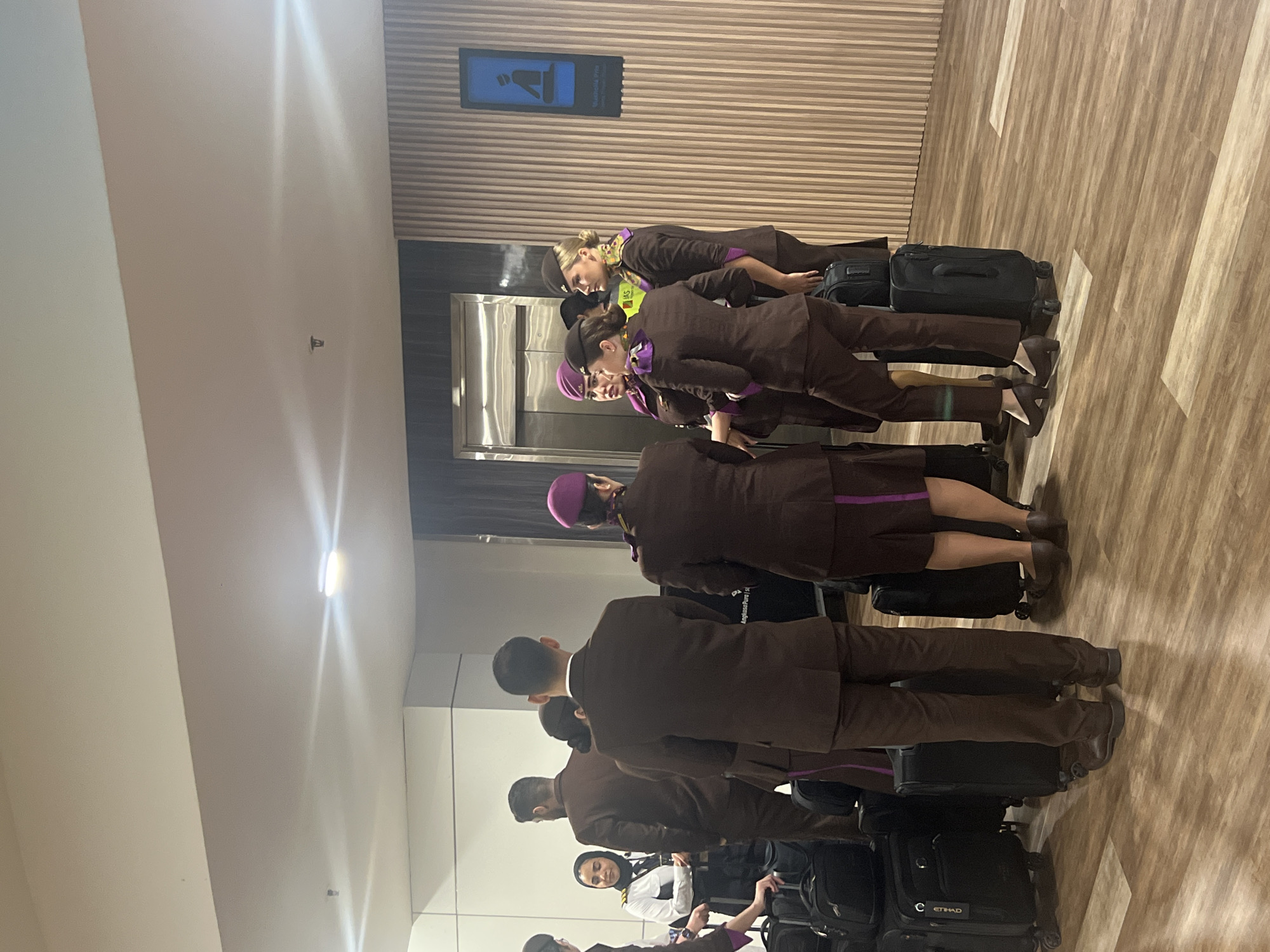 A group of airline crew members stands in line with their luggage in an indoor location. The crew members are wearing brown uniforms with purple accents. The luggage consists of black rolling suitcases, some with "Etihad" labels. The location appears to be an airport terminal, evidenced by a sign with an image of a seated figure, possibly indicating a waiting area. The floor has a light brown wood-like pattern. The ceiling is white with built-in lighting. The crew is near a metal elevator.
While the image suggests an airport setting, and Etihad Airways operates out of multiple international locations, there is no visible information to definitively confirm the location as Kuta, Indonesia.
