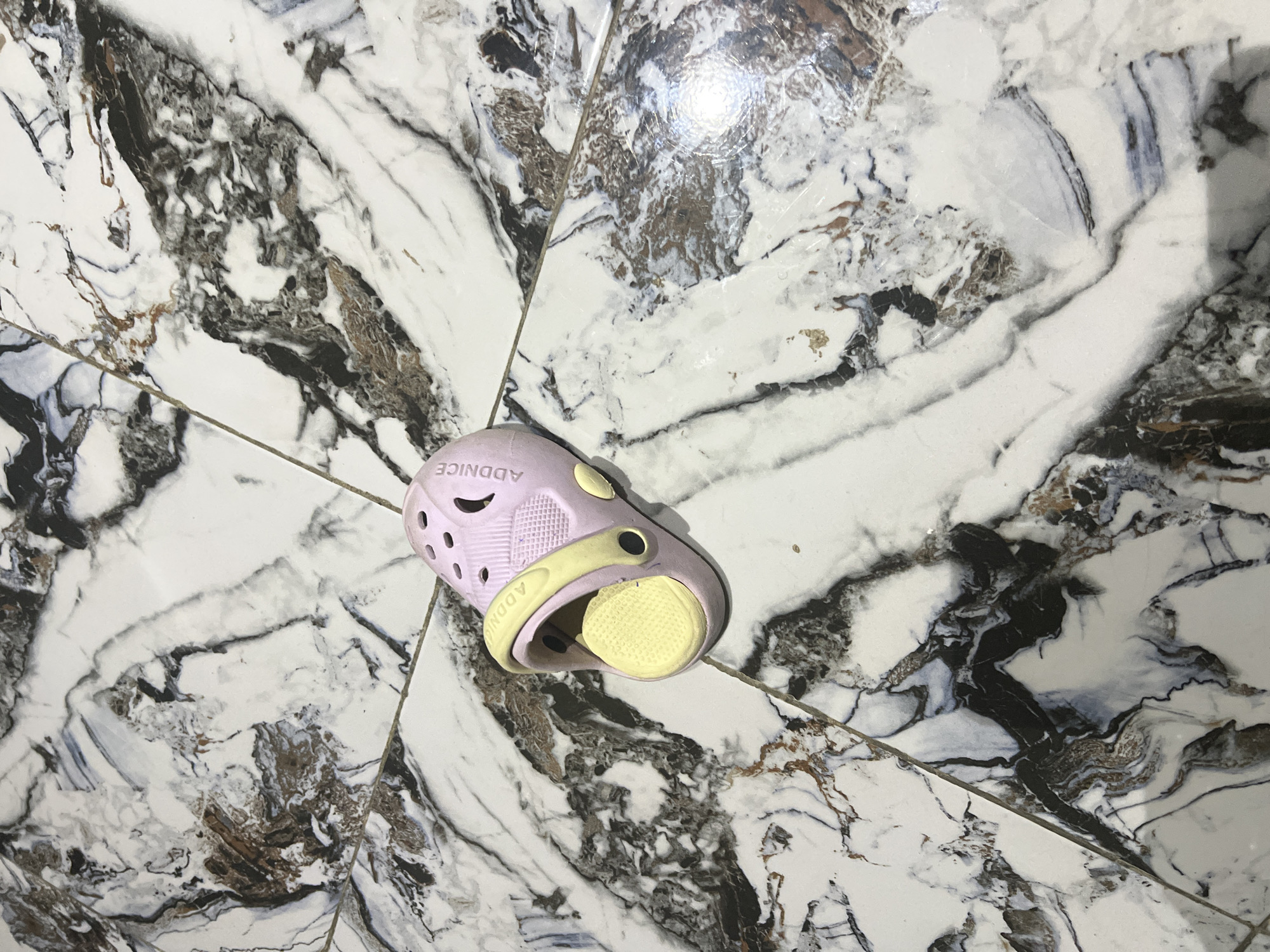 A single pink and yellow child's clog is pictured on a marble-patterned tiled floor. The clog is lying on its side with the opening facing upwards, revealing a yellow sole insert. The clog has ventilation holes on the top and the word "ADDNICE" is embossed on the side. The floor tiles are predominantly white with black and brown veining. The lighting suggests an indoor environment, possibly a home, and the scene is static. The image does not contain any information that would allow for a specific location within Jalingo, Nigeria to be determined.