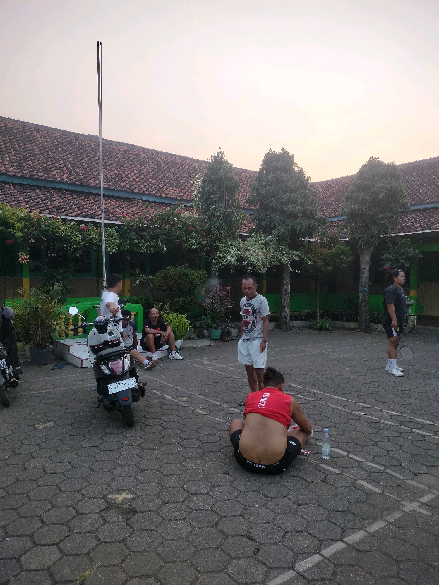 The image depicts an outdoor scene, likely a schoolyard or public court, in Boja, Indonesia, during what appears to be late afternoon or early morning, given the soft lighting. Several men are present, with some engaged in or preparing for a game of badminton.
In the foreground, a man in a red tank top and black shorts is sitting on the hexagonal paved ground, facing away from the camera, with his back partially exposed. A water bottle and what appears to be a pack of cigarettes are beside him.
To his left, a dark-colored scooter is parked. In the mid-ground, three men are standing. One man in a white t-shirt and shorts is standing near the scooter. Two other men are standing in what looks like a badminton court area. One, wearing a gray t-shirt and white shorts, appears to be talking. The other, wearing a dark t-shirt and shorts, is holding a badminton racket. Further to the right, another man in a dark t-shirt and black shorts is standing, also holding a badminton racket.
In the background, a building with a red tile roof and green trim is visible, with trees and foliage surrounding it. A tall flagpole stands near the building. The ground is paved with interlocking hexagonal blocks. The overall impression is a casual gathering for sport and relaxation.