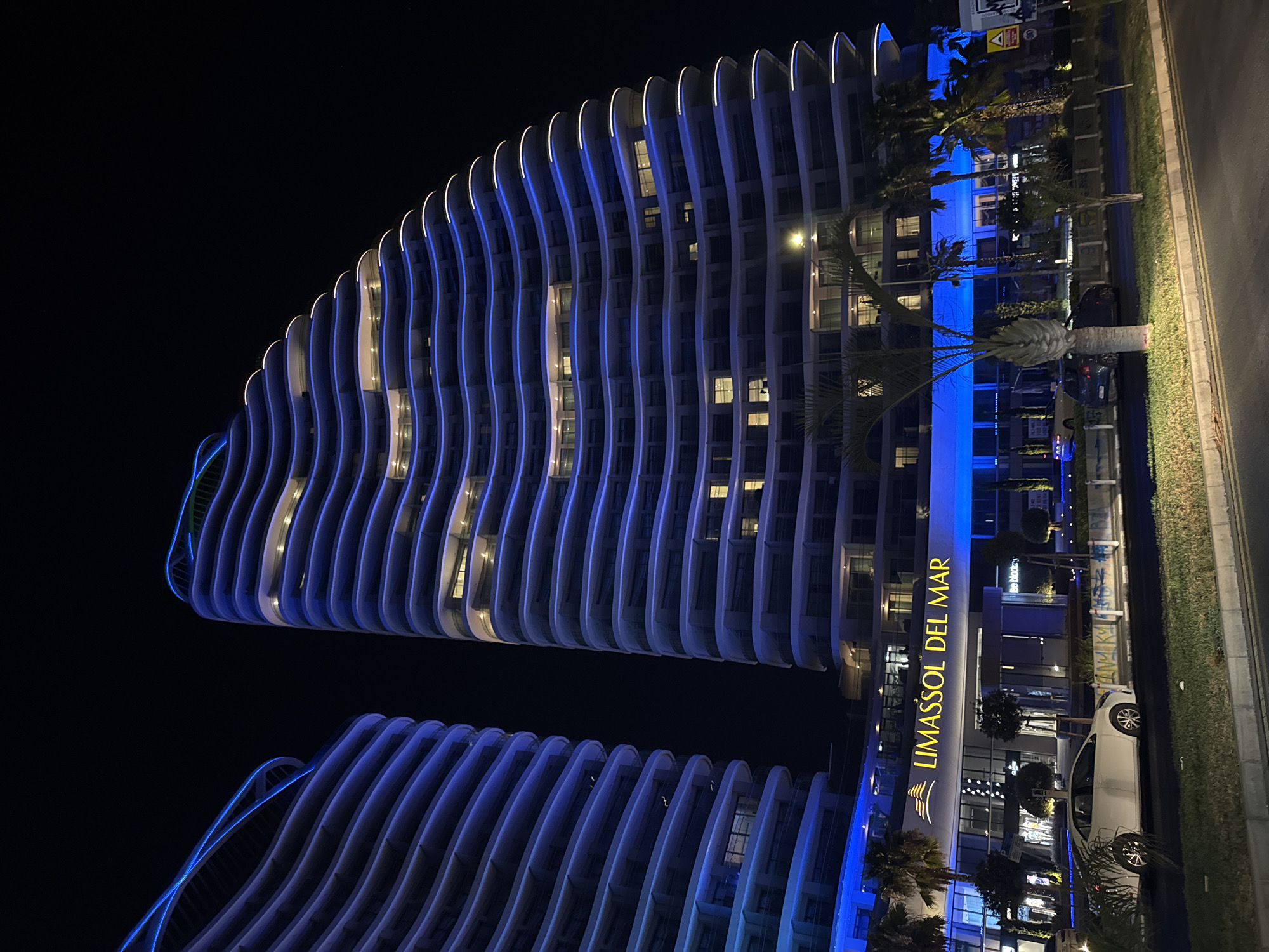 In Germasogeia Municipality, Cyprus, at night, two modern, wavy-shaped skyscrapers are illuminated with blue and white lights. The skyscraper on the right is taller and wider, with undulating balconies. The skyscraper on the left is partially visible and also features a wavy design. Below the skyscrapers, a building with the illuminated text "LIMASSOL DEL MAR" is visible. In front of the buildings, a white car is parked on the street. Palm trees and other greenery are present along the sidewalk and in front of the buildings. The scene is lit by the building's lights and streetlights, casting a nighttime ambiance.