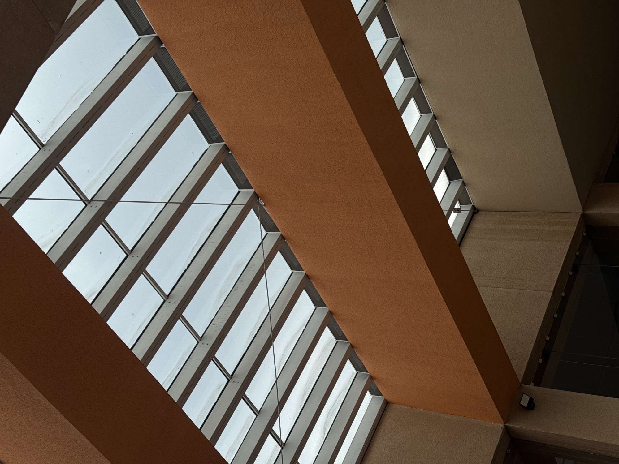 The image is an upward-facing shot of a building's interior architecture, specifically a portion of the roof or ceiling. The scene is characterized by a grid-like structure of glass panes separated by metal or concrete beams, creating geometric patterns. The light coming through the glass suggests it is daytime, with a diffuse or overcast sky visible outside. A broad, textured beam of brownish-orange hue crosses the frame diagonally, partly obscuring the view of the glass structure. Other portions of the ceiling or roof are visible in various shades of brown and beige, indicating different sections or levels of the structure. The architecture has a modernist or brutalist style.
Based on the architecture and geometric designs, this image is very likely the Papalote Museo del Niño in Mexico City, Mexico.