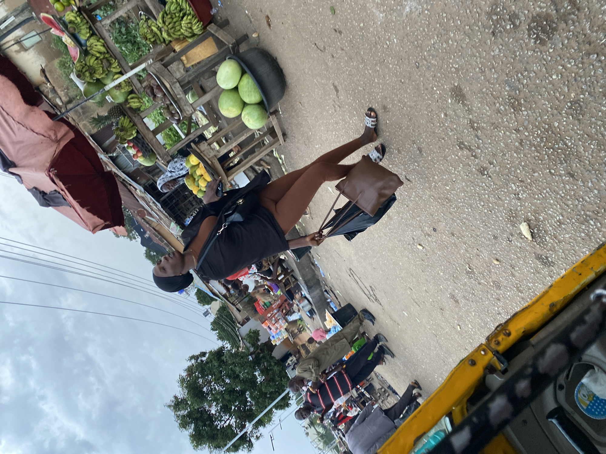The image depicts an outdoor scene, possibly a market, in the city of Bukuru, Nigeria, under an overcast sky. A woman in a black headscarf and black abaya, wearing brown leggings and sandals, is the central figure, walking away from the camera and holding two bags. She is seen in front of a fruit stand that displays watermelons, bananas, pineapples, and other produce. Several other people are visible in the background, some standing and others walking. There is a yellow vehicle in the foreground, partially obscuring the lower left of the frame. The ground is asphalt, with some debris scattered across it. The overall impression is of a bustling street scene.