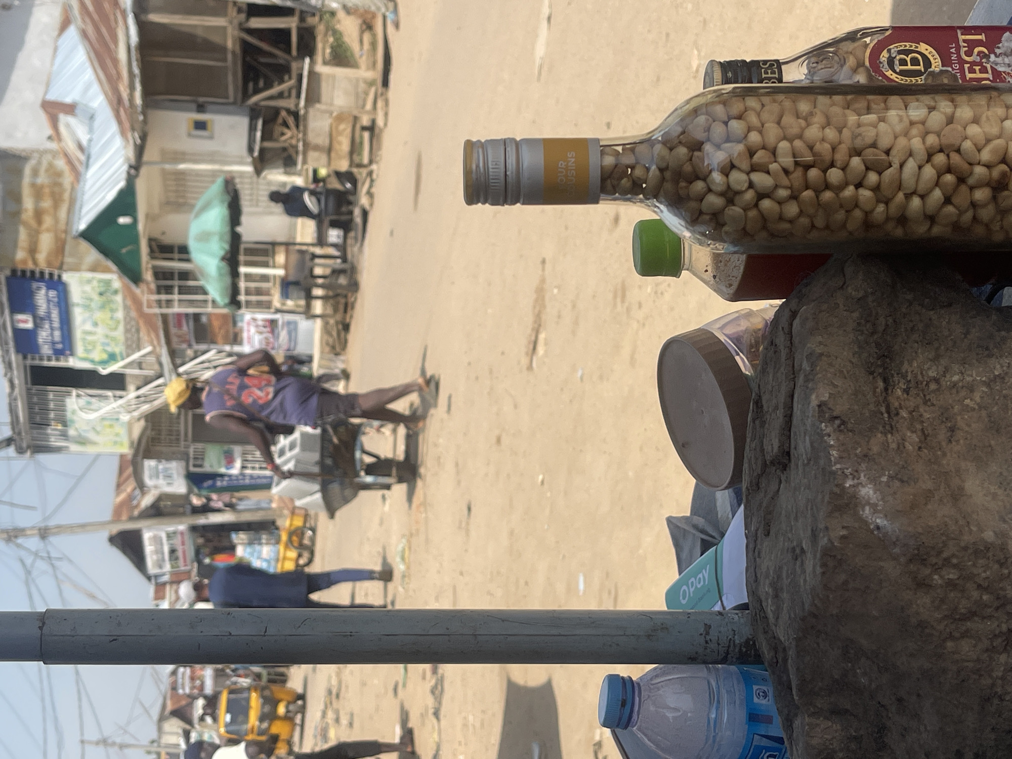 In a dusty street market scene in Abuja, Nigeria, a man wearing a yellow cap and a purple sleeveless jersey with the number 24 on it is pushing a wheelbarrow. He is walking away from the camera, towards the right side of the frame. In the foreground, a large rock sits on a table, with a bottle of water, a container with a brown lid, and two glass bottles filled with nuts and liquid placed on it. The bottle in the center is a clear glass bottle filled with peanuts. Behind the man and wheelbarrow, a yellow tricycle is visible, as well as various market stalls with corrugated metal roofs and awnings. People are seen walking and sitting in the background. The ground is sandy and uneven, and power lines are strung overhead. The overall impression is of a bustling, informal market environment.